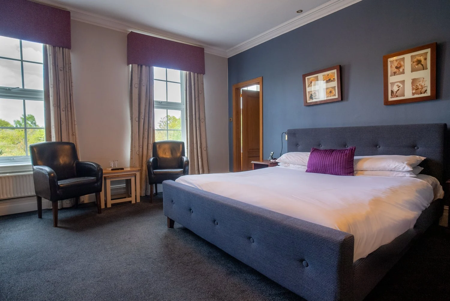 Hotel bedroom with a large bed, two black armchairs, a small wooden table, two windows with curtains, framed wall art, and a nightstand with a phone and lamp.
