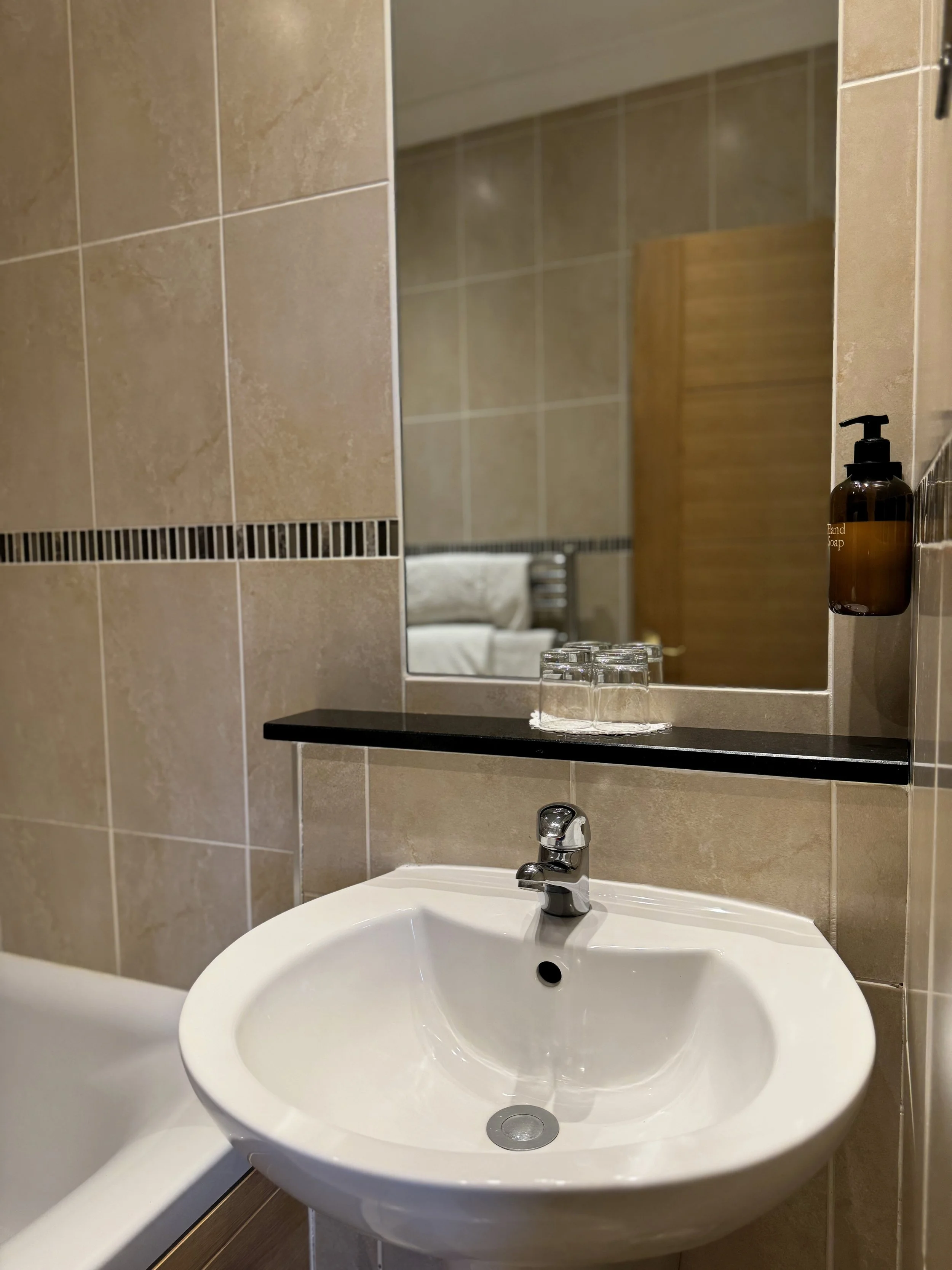 A bathroom sink with a mirror above it, glass cups on a black shelf, and soap dispenser on the wall to the right.