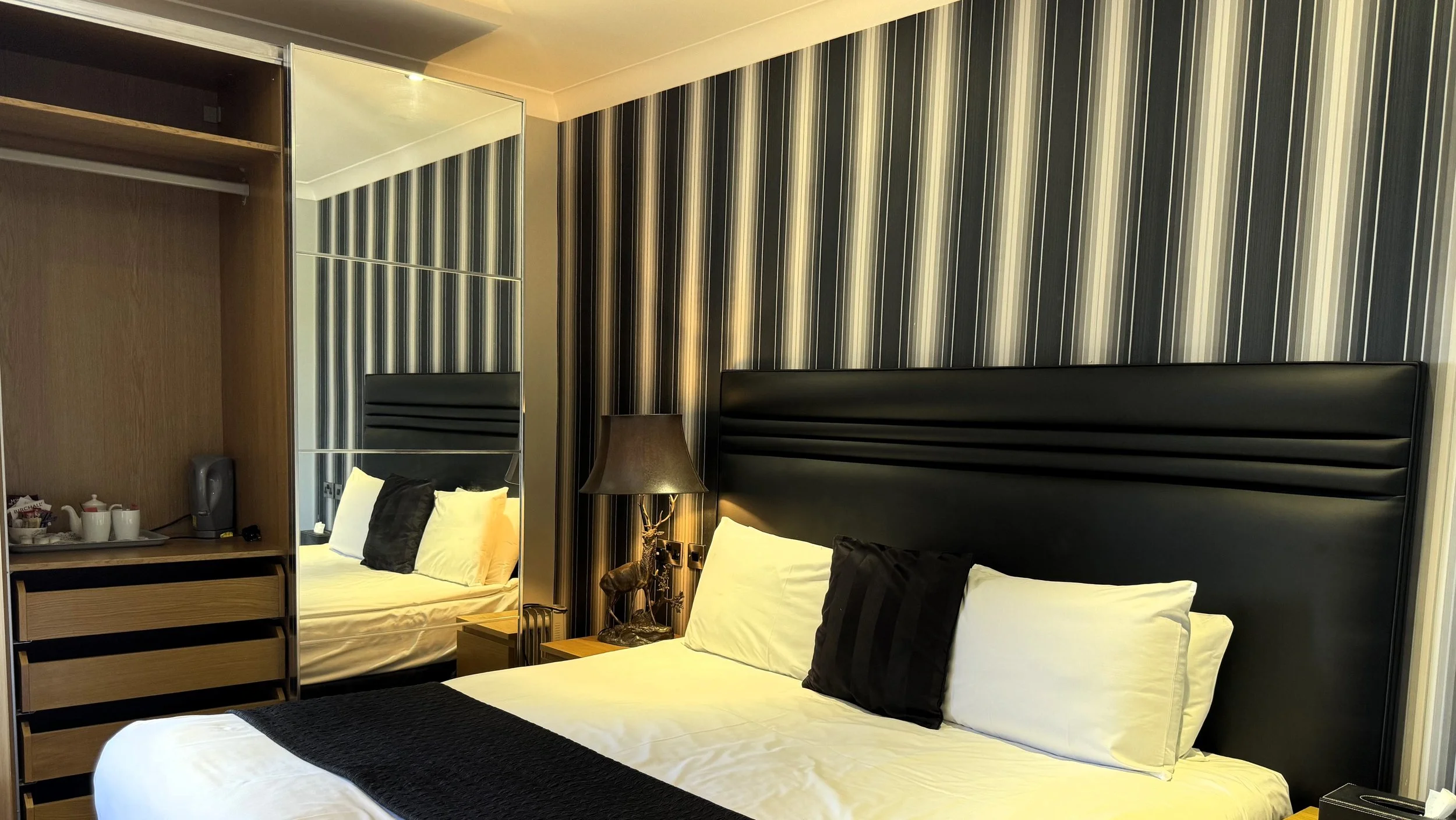 A hotel bedroom with a large bed, black headboard, black and white bed linen, and black and white striped wallpaper. A bedside table with a lamp and decorative reindeer sculpture, a mirrored wardrobe, and a small bar area with tea and coffee supplies