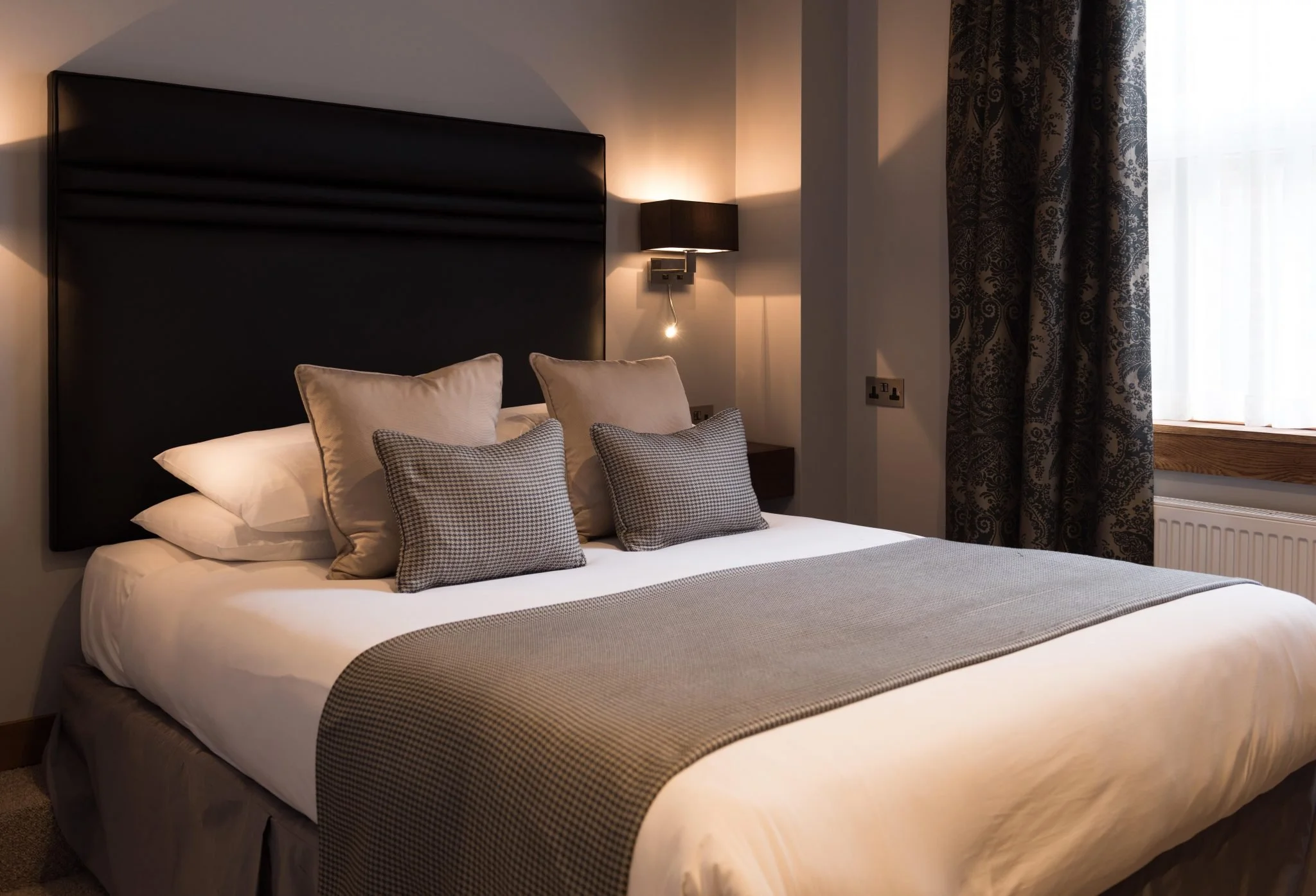 A neatly made bed with beige and gray pillows, a gray blanket, and a dark headboard in a cozy hotel room with a window and curtains.