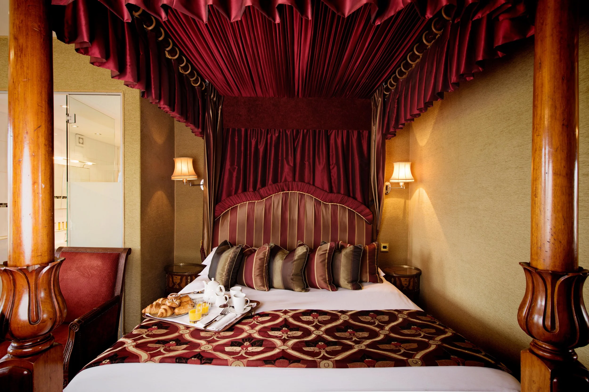 A luxurious hotel room with a four-poster canopy bed with deep red curtains, decorative pillows, and a breakfast tray with croissants, juice, coffee, and utensils.