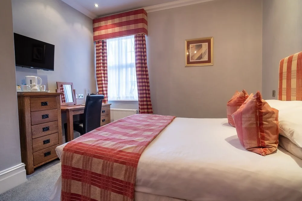 Hotel bedroom with a bed, pillows, window with red checkered curtains, wooden desk, chair, TV, and framed artwork.