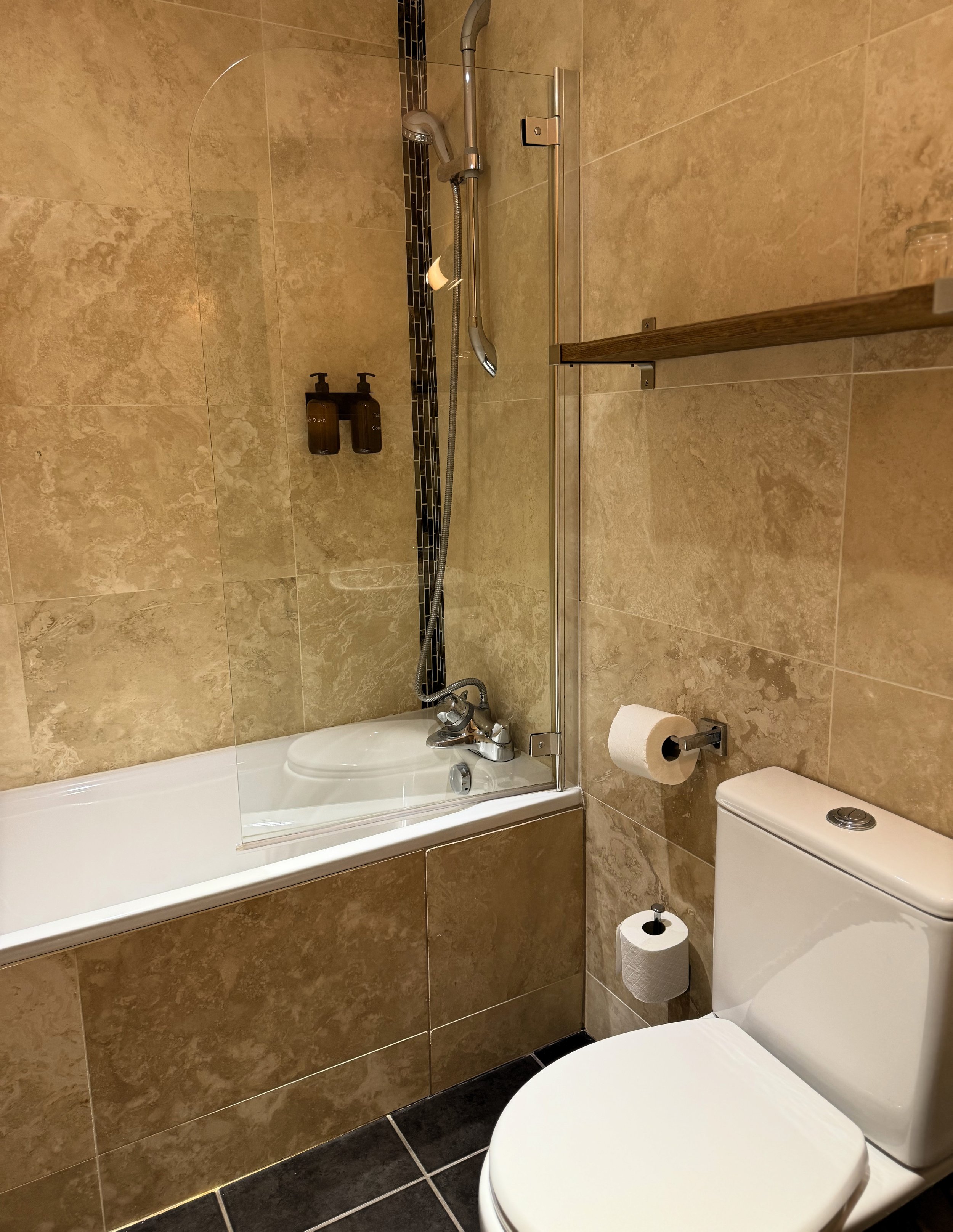 Bathroom with a bathtub, showerhead, two brown soap dispensers, toilet paper roll, and toilet, with beige tiled walls and dark tiled floor.