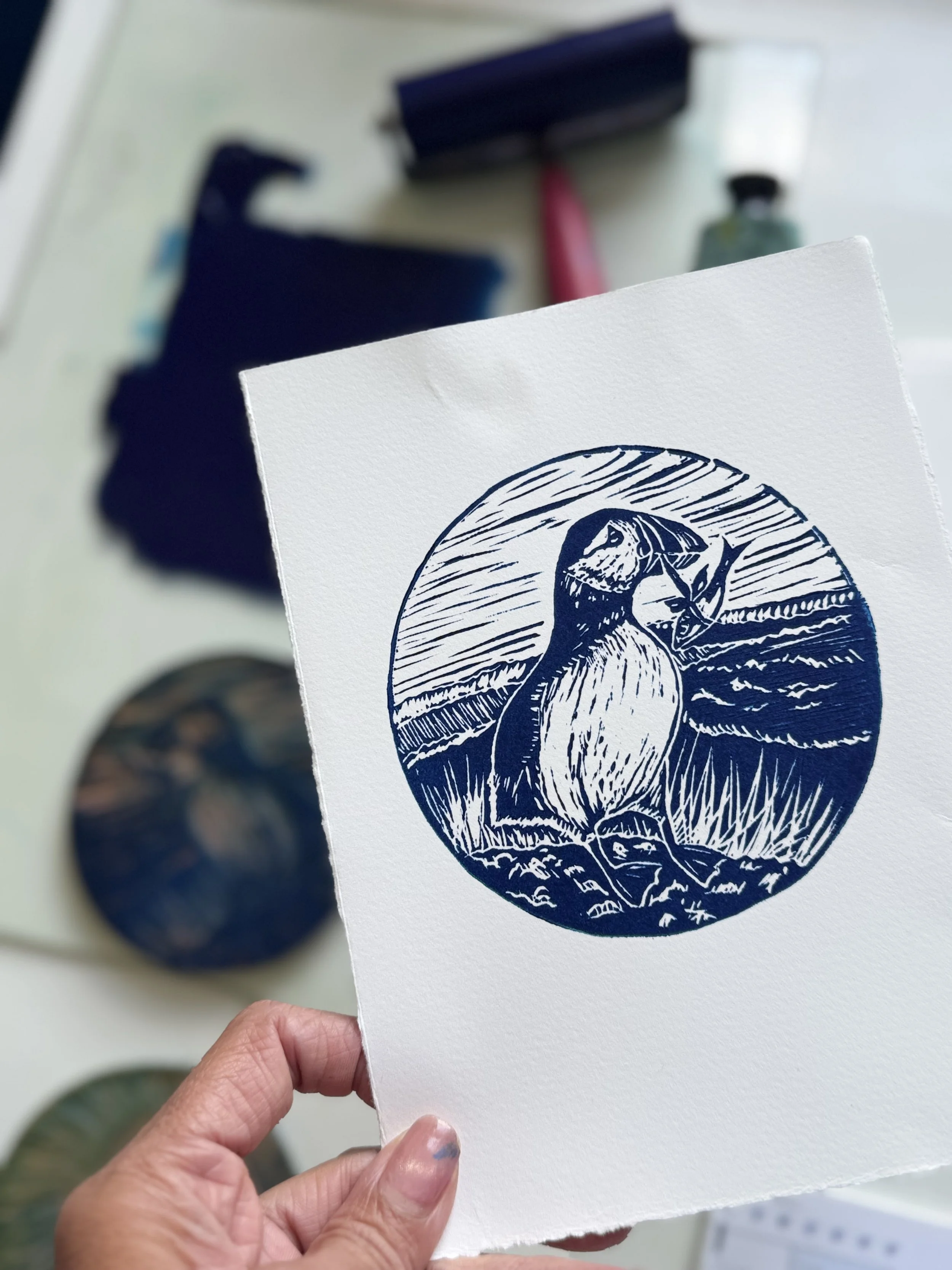 Print of a blue and white illustration of a bird, possibly a puffin, holding a branch, with a landscape background.
