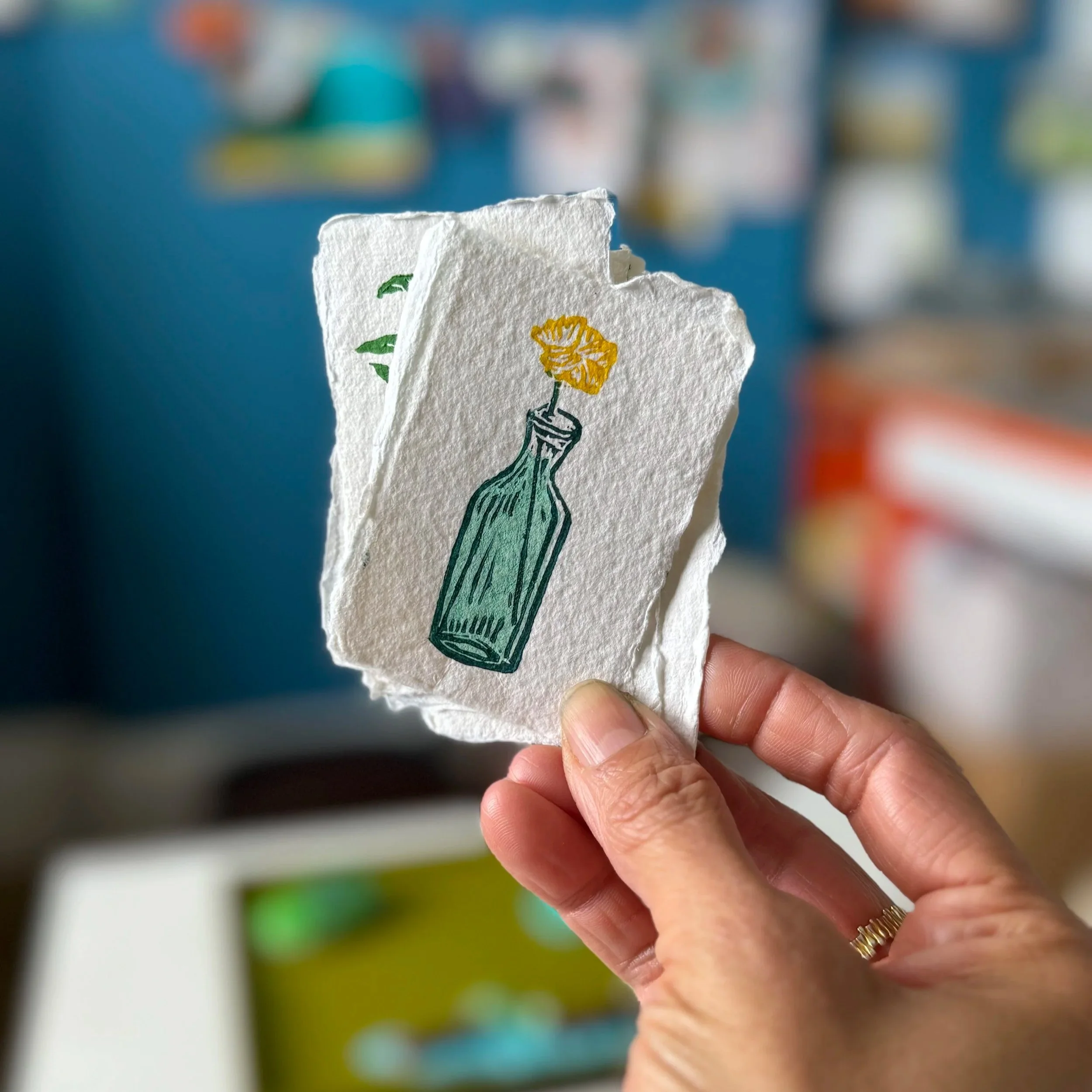 Close-up of a person's hand holding a rough-edged piece of paper with a hand-drawn illustration of a green bottle with a yellow flower on top, with a blurred background.