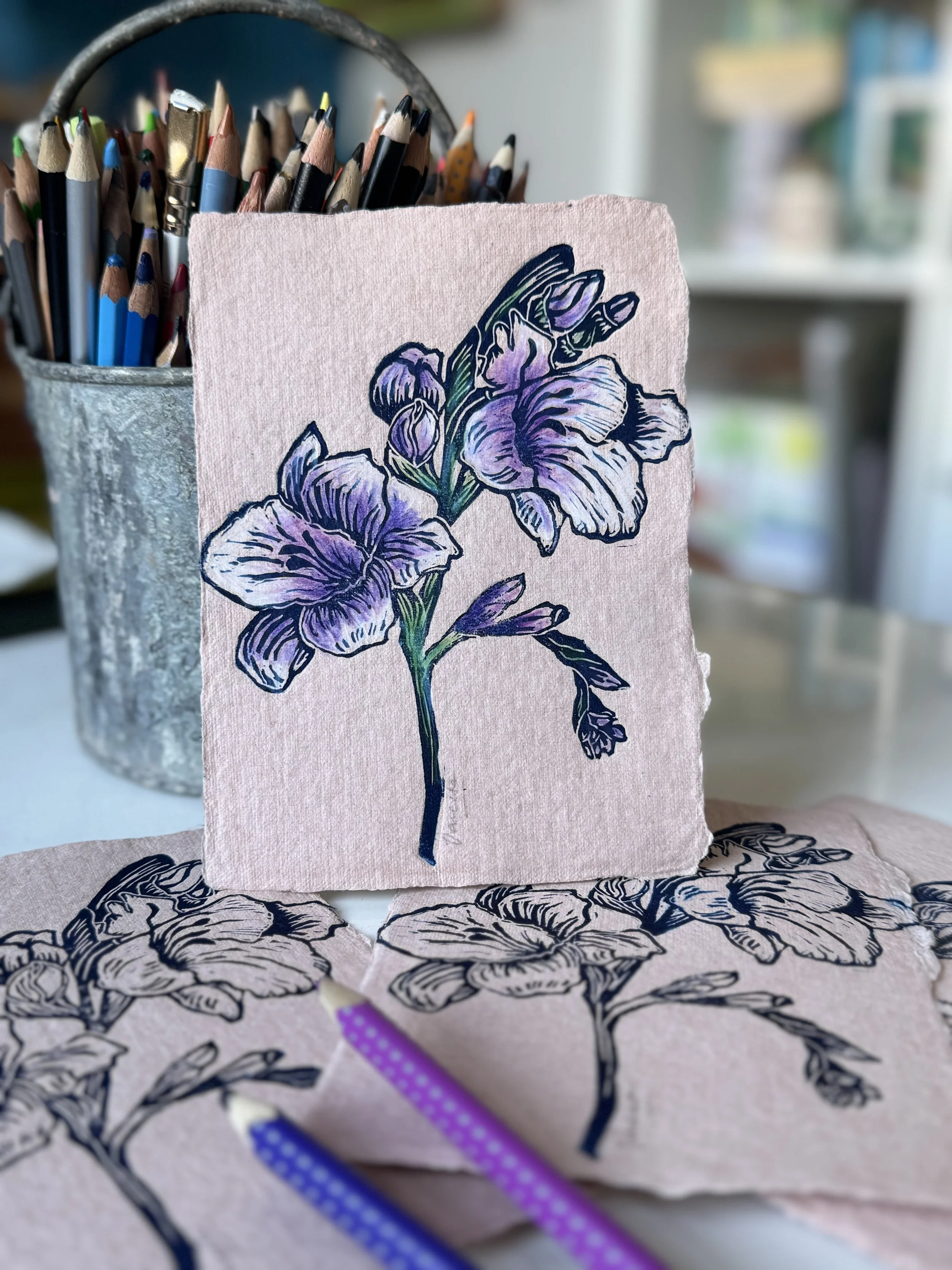 Picasso-style drawing of a purple iris flower on textured pink paper, with colored pencils and drawings of the same flower in the foreground, and a cup of pencil holders in the background.