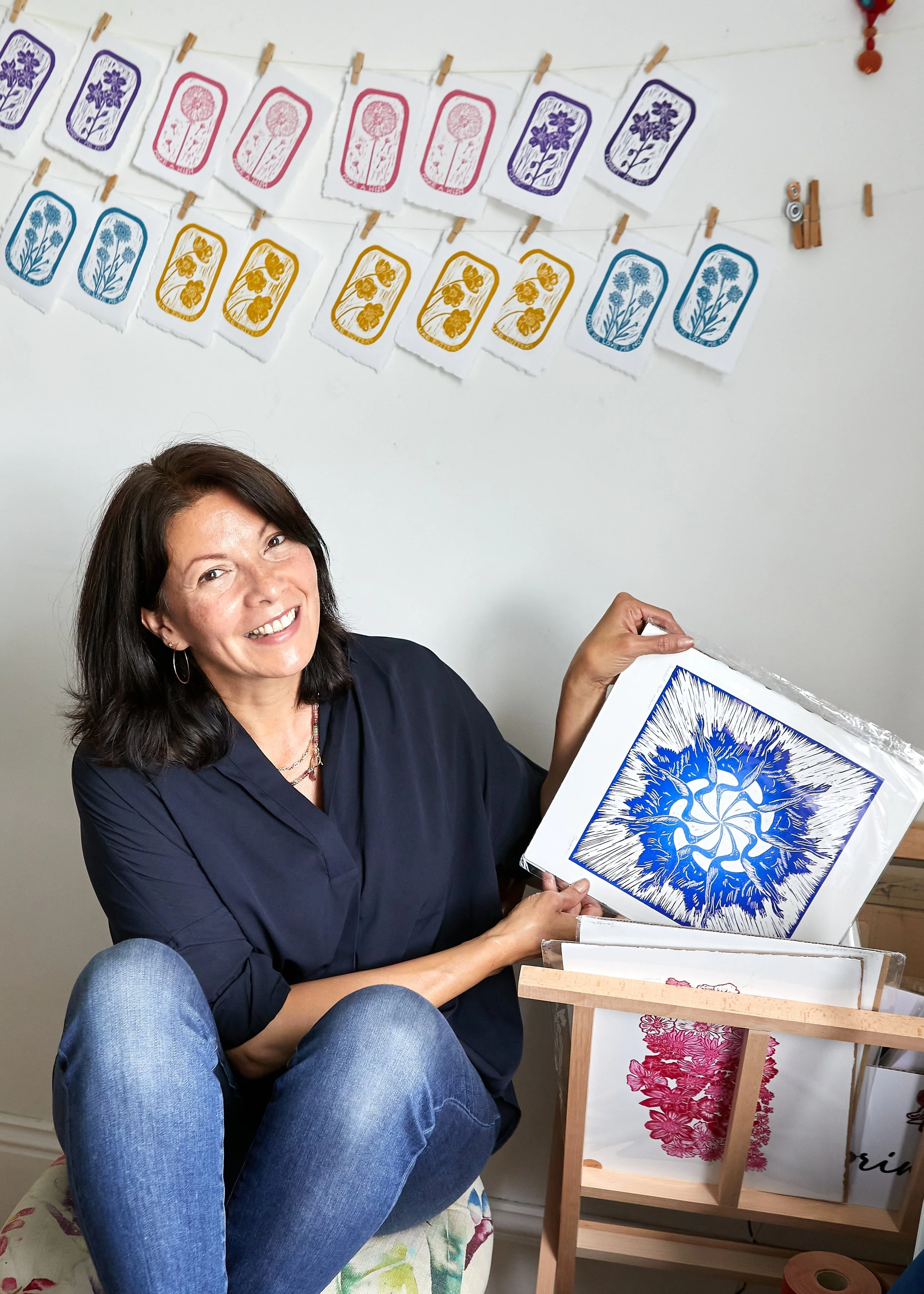 A woman sitting on a floral cushion, smiling, holding a framed piece of art with a blue and white abstract floral pattern. Behind her, there are paper prints with floral designs in multiple colors hanging on a white wall.