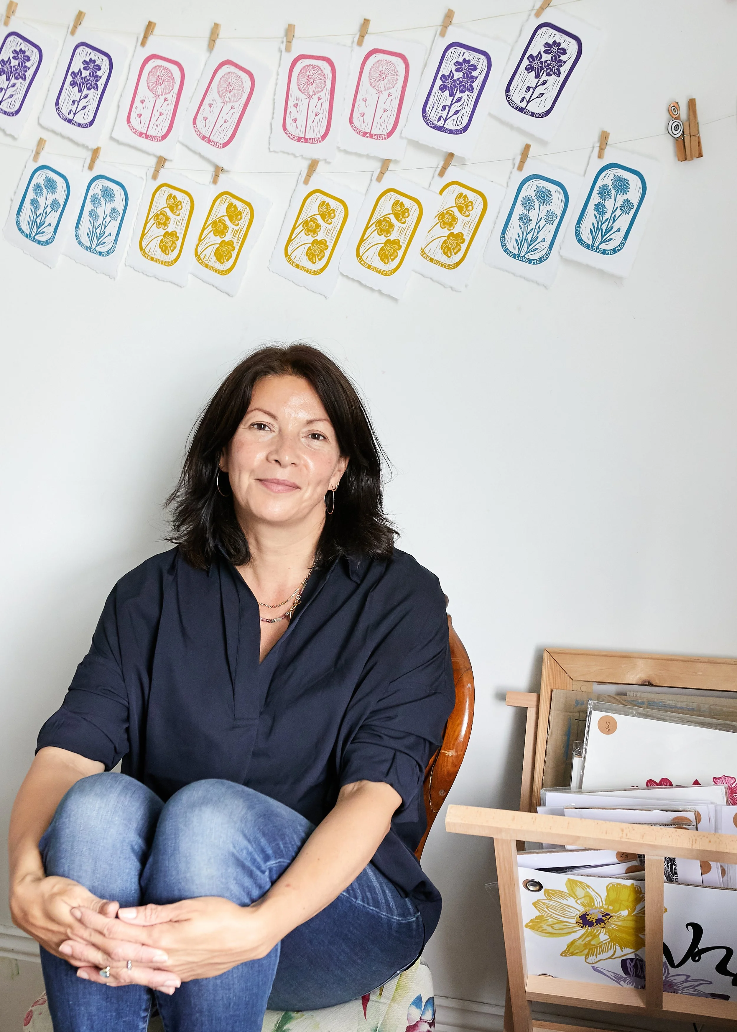 A woman with shoulder-length dark hair wearing a black blouse and blue jeans, sitting with her knees pulled up to her chest in a room with decorative paper banners of colorful floral and nature designs hanging on the wall behind her, and a wooden magazine or photo display stand with various papers and artwork.