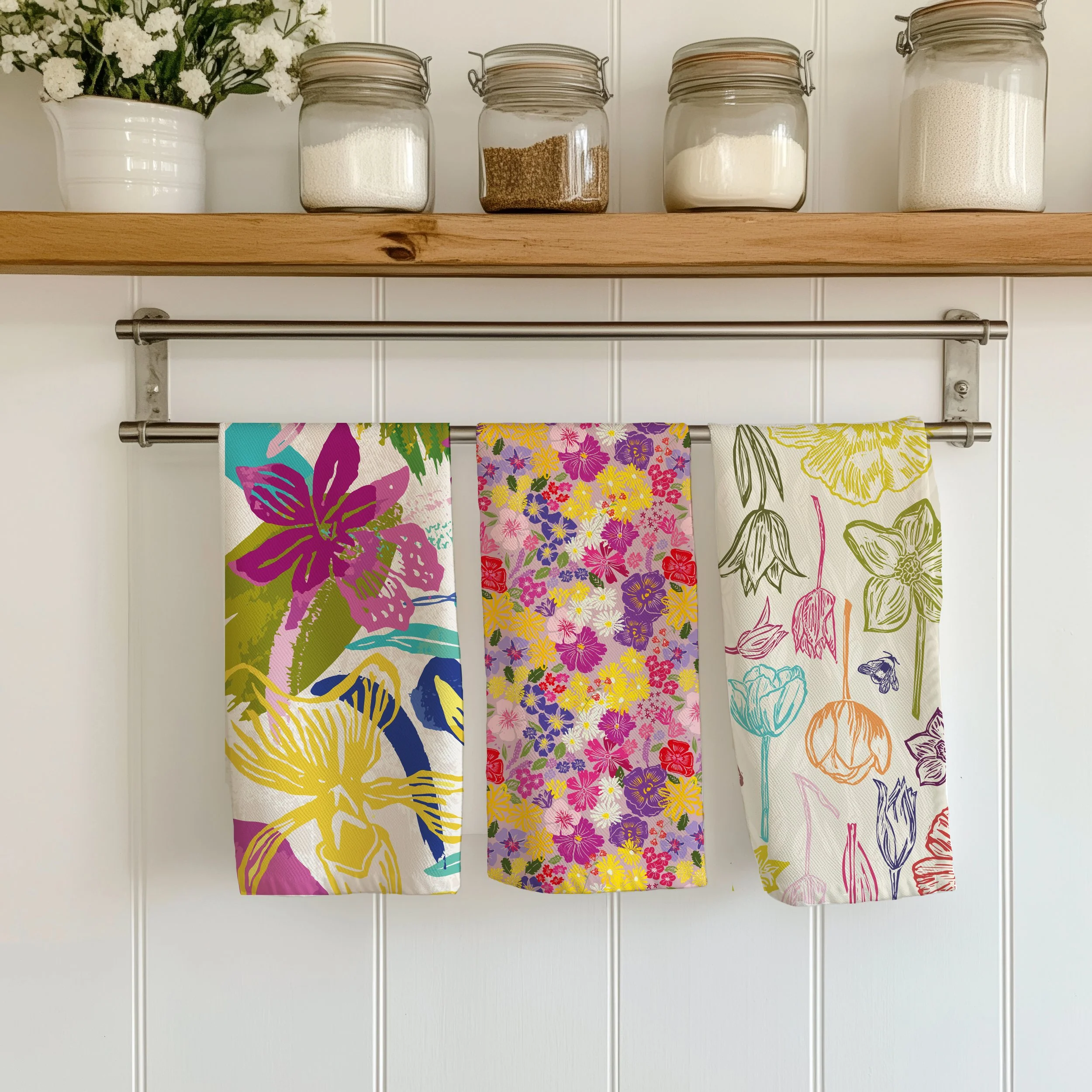 Three colorful floral patterned towels hanging on a metal rod in front of a white wall with a wooden shelf holding glass jars filled with different types of sugar and a flowerpot with white flowers.