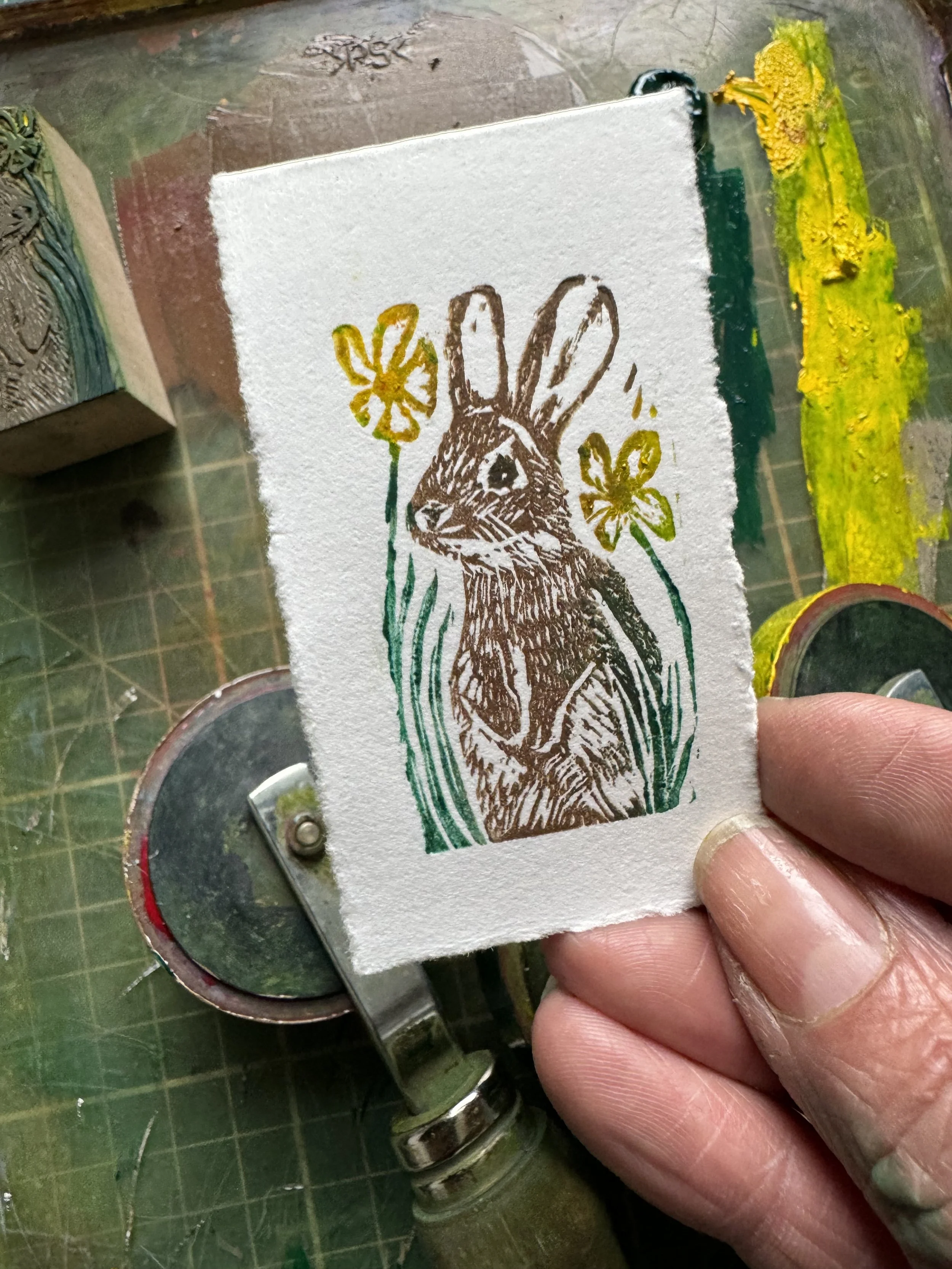 A hand holding a piece of paper with a stamped and colored illustration of a rabbit, surrounded by flowers and grass, on a woodworking or stamping workstation.