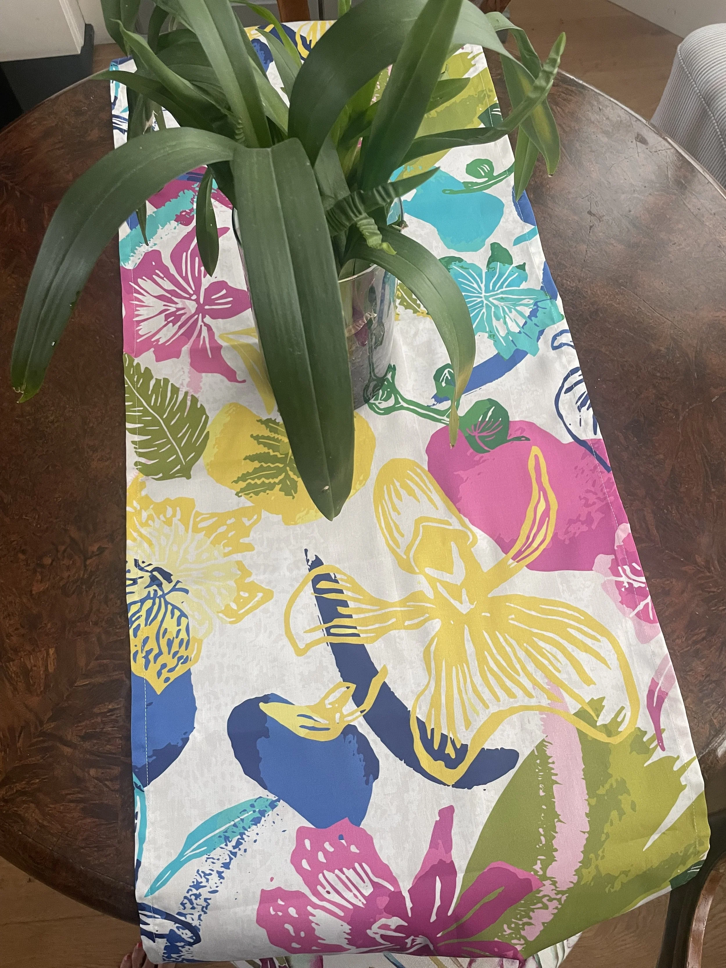 A table runner with a colorful floral and butterfly pattern in pink, yellow, blue, green, and white, placed on a wooden table, with a green potted plant on top.