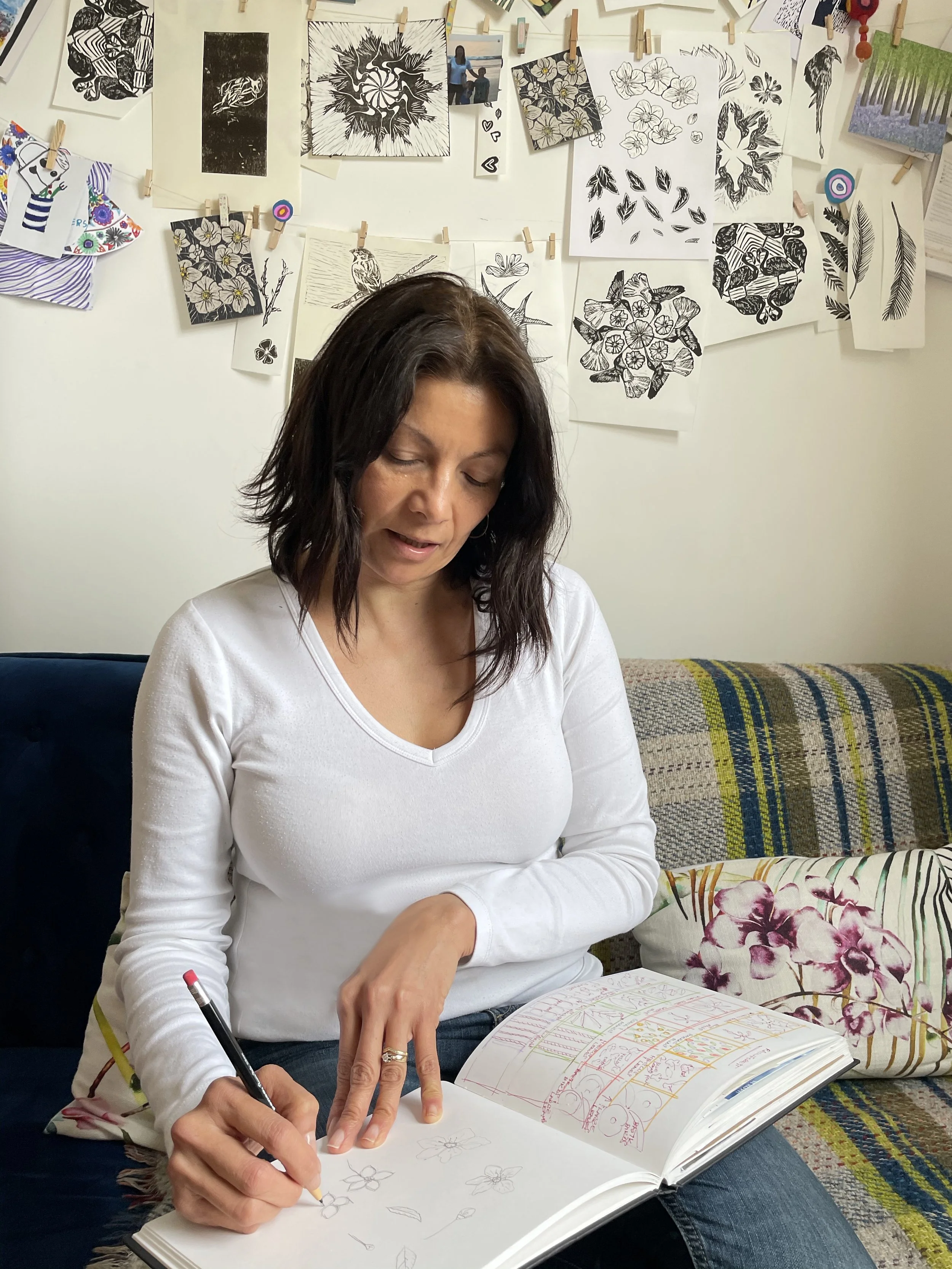 A woman with dark hair in a white long sleeve shirt sitting on a couch drawing flowers in a sketchbook.