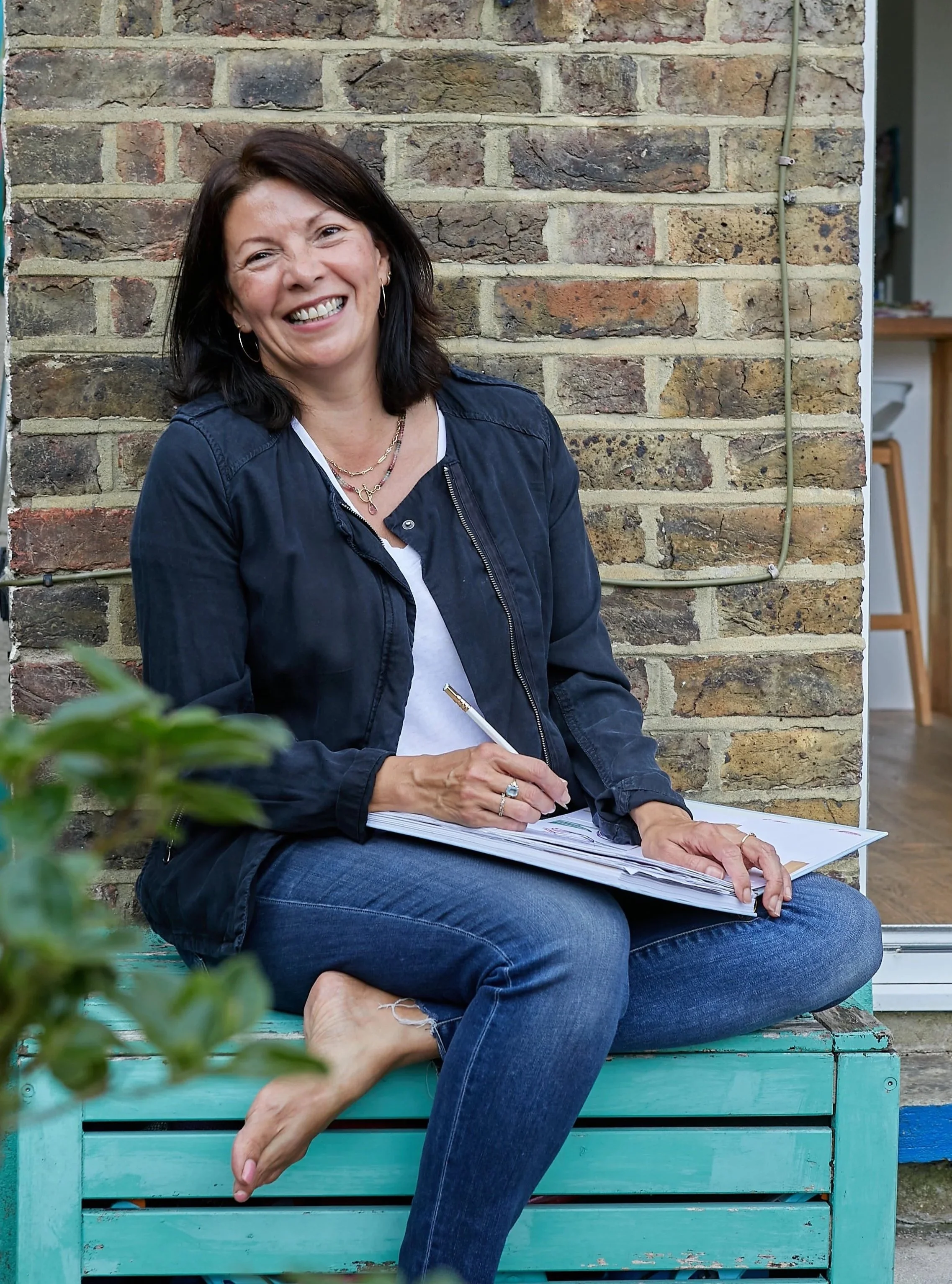 A woman with dark hair, smiling, sitting cross-legged on a turquoise wooden bench in front of a brick wall, holding a pen and a notebook. She's wearing a black jacket, white shirt, and blue jeans.
