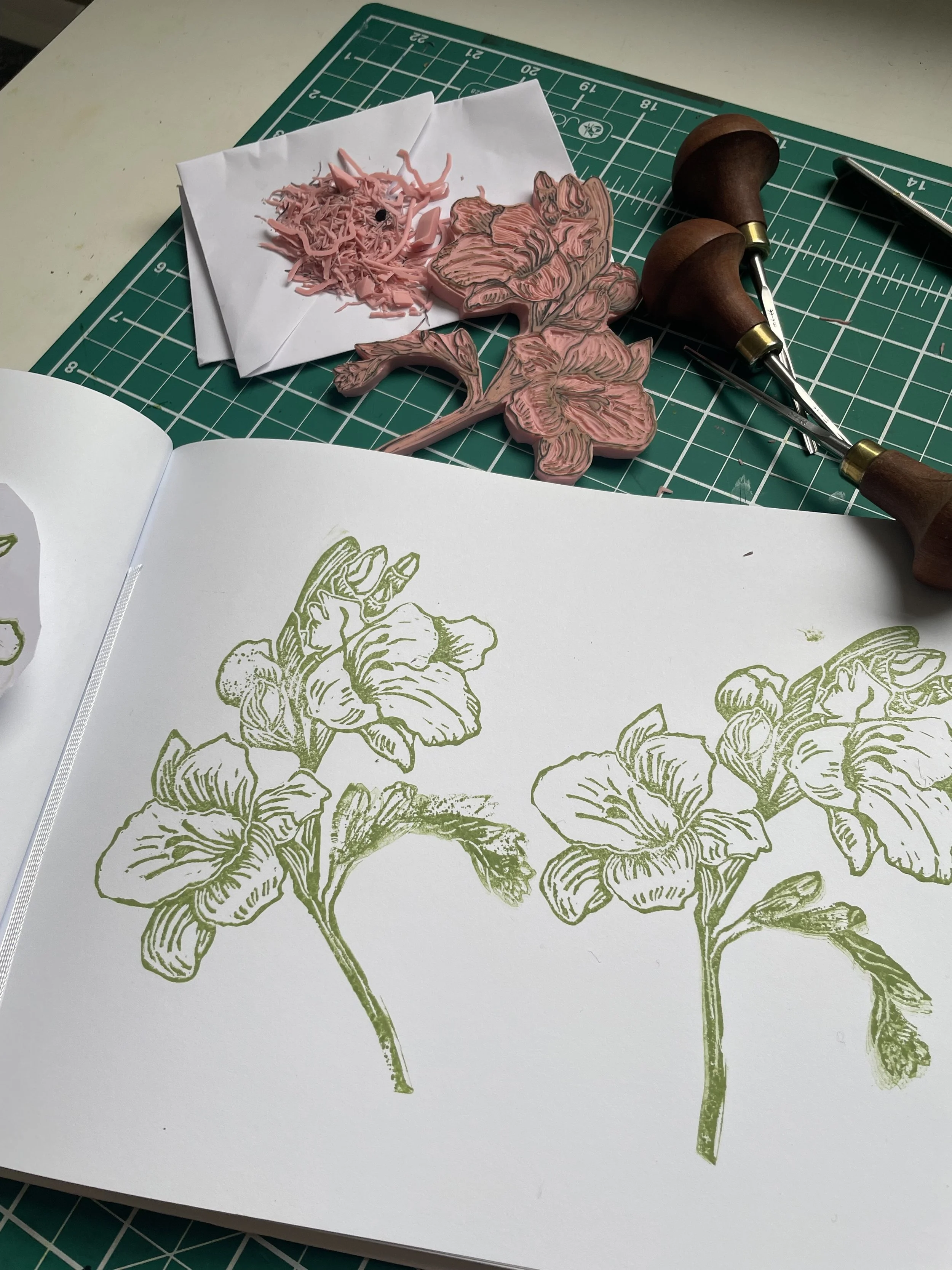 A workspace with a botanical art print of flowers on white paper, rubber stamps of the same floral design, ink pads, and carving tools, all on a green cutting mat.