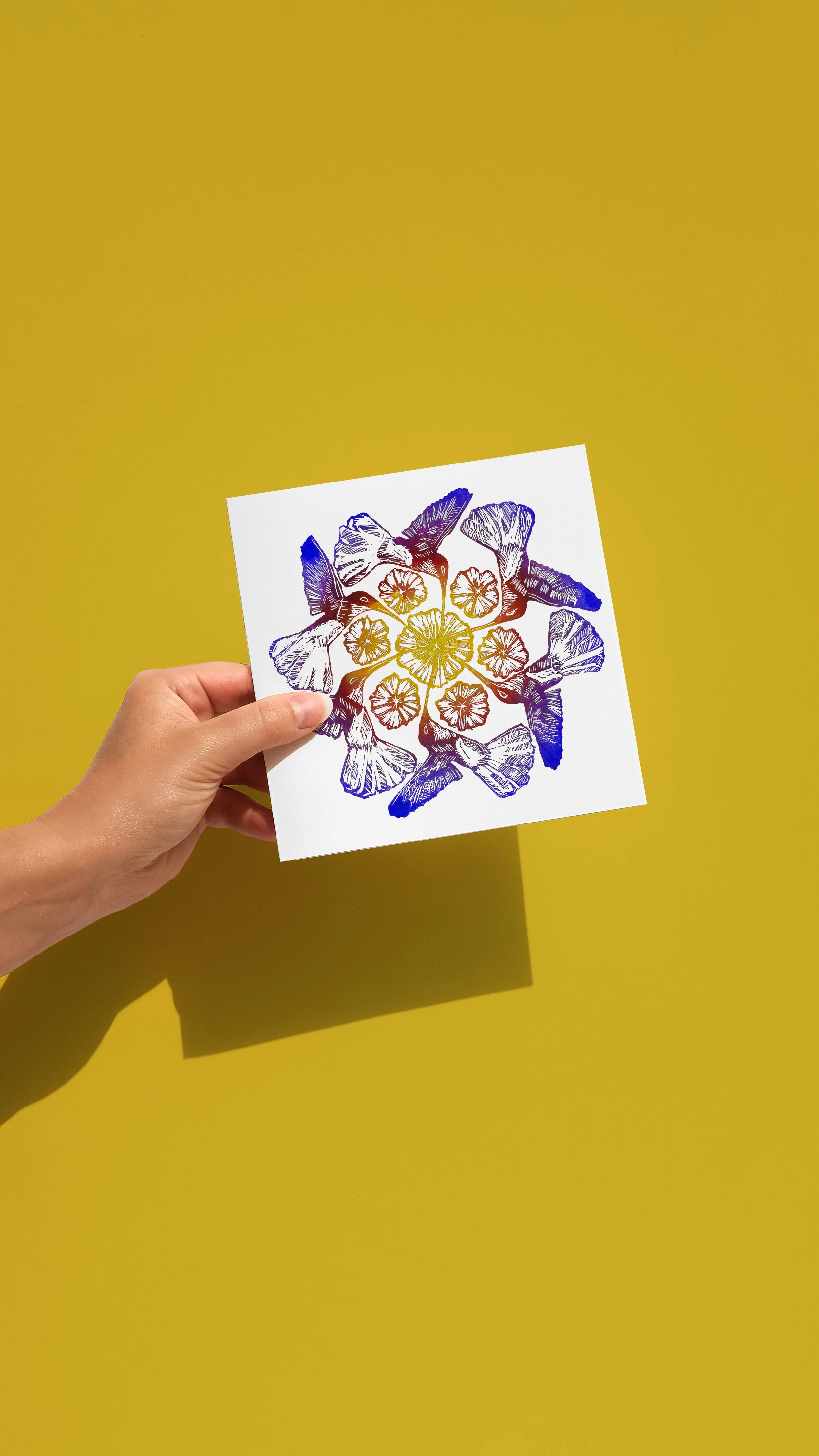 Lino print design on a card featuring 6 Jacobin hummingbirds fluttering around 7 peony flowers. Printed in bright ombre shades of ultramarine, deep red and golden yellow