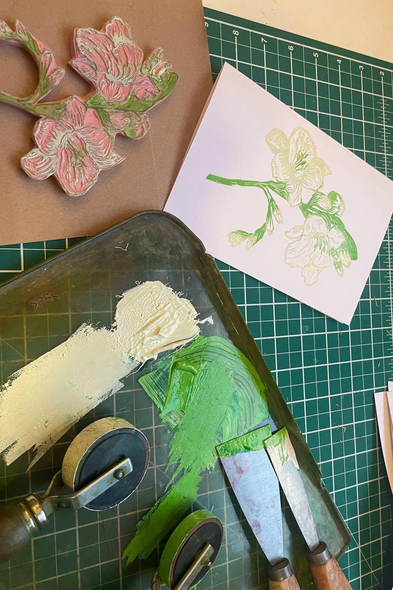 A worktable with art supplies showing a pink and green floral wood cutout, a painted floral illustration on paper, and a tray with white and green paint along with painting knives.