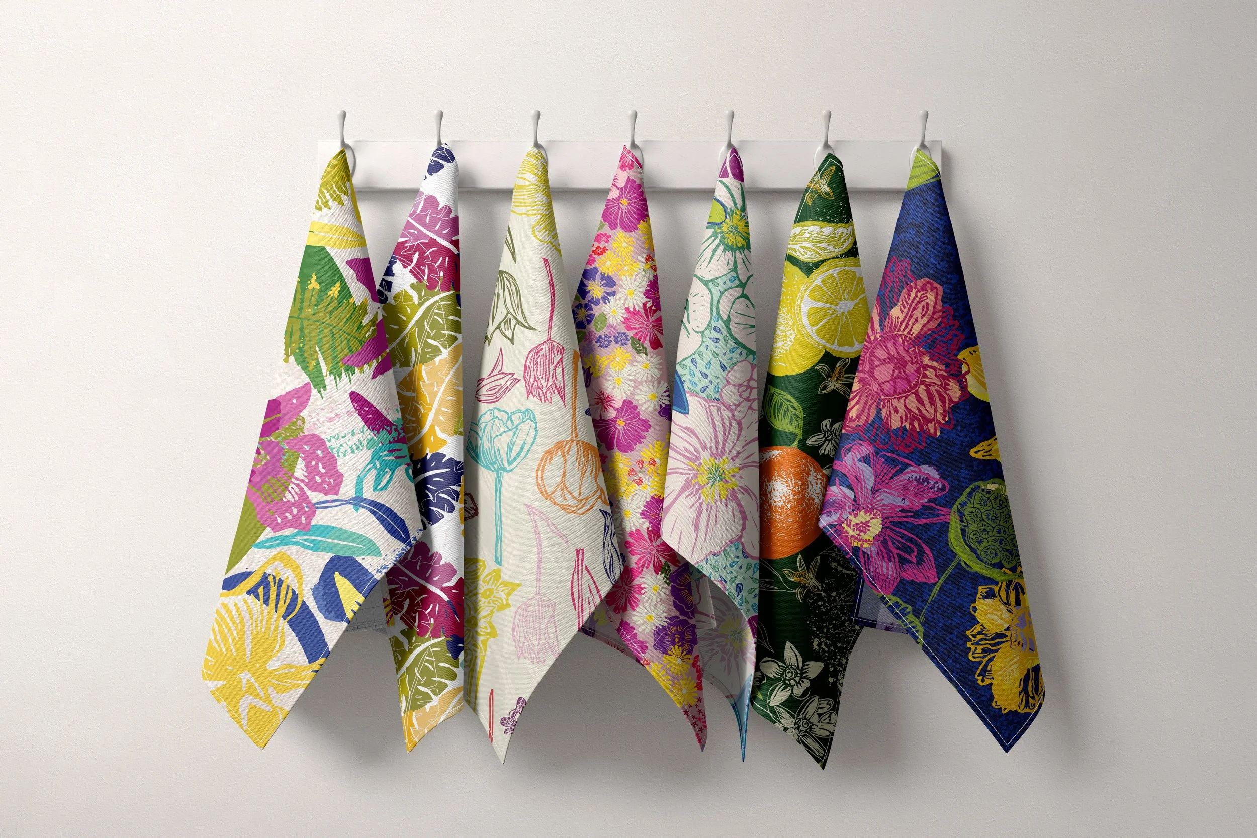 Six colorful printed dish towels hanging on white hooks against a plain wall.