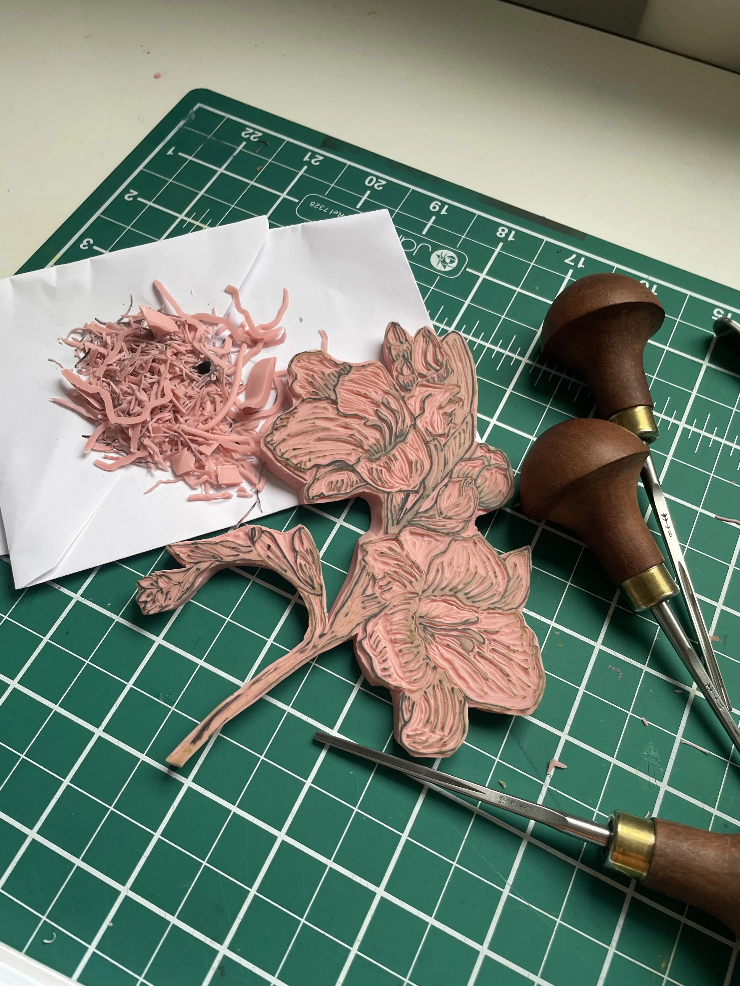 A floral rubber stamp with pink and gray details, wood-handled carving tools, a paper with pink shredded rubber, and a cut-out paper flower stamp on a green cutting mat.