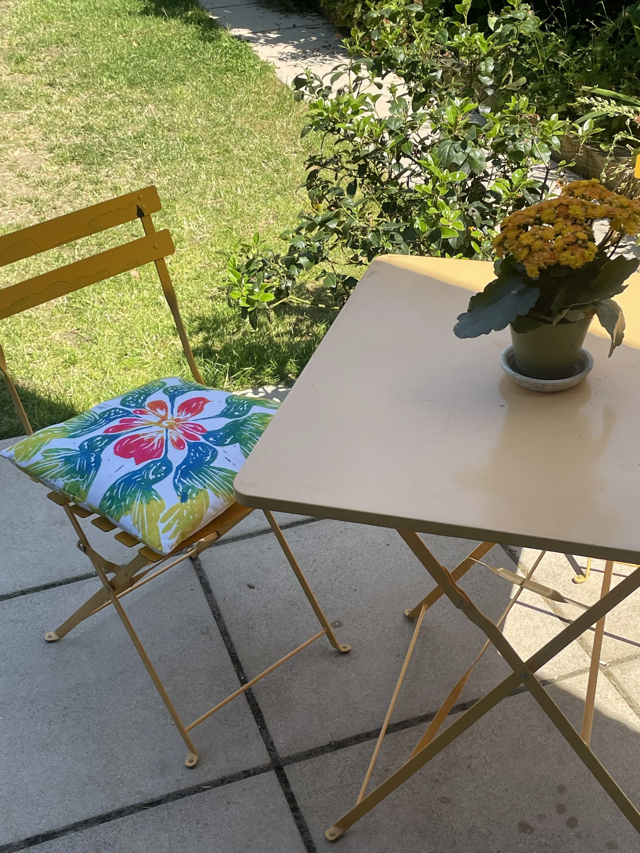 Small outdoor patio table with a potted yellow flowering plant, a mustard yellow chair with a colorful floral cushion, green grass, bushes, and a walkway in the background.