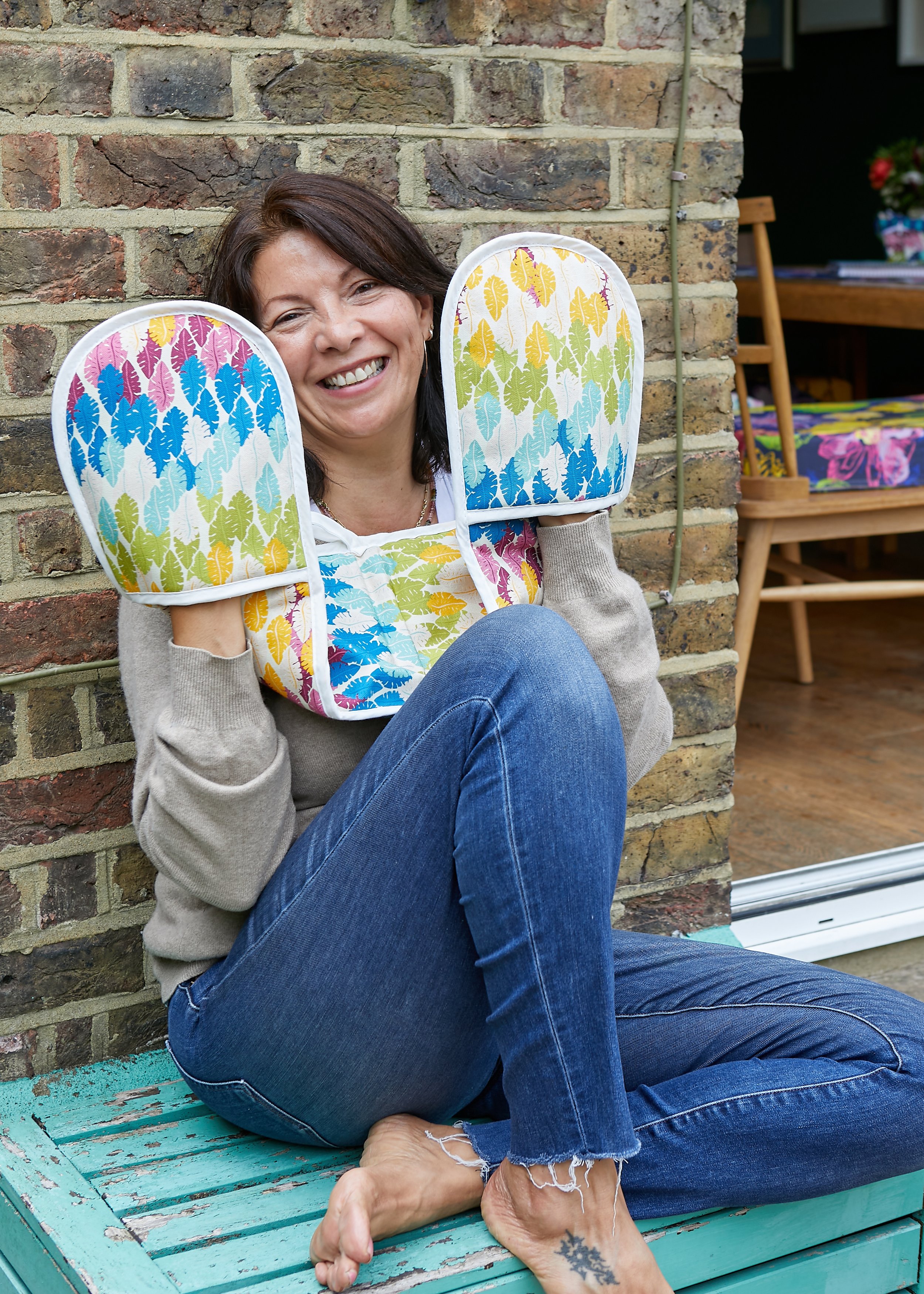 A woman smiling while sitting on a turquoise wooden bench outdoors, holding colorful oven mitts with leaf patterns. She is wearing a beige sweater and blue jeans, with a tattoo visible on her ankle. There is a brick wall behind her and garden furniture in the background.