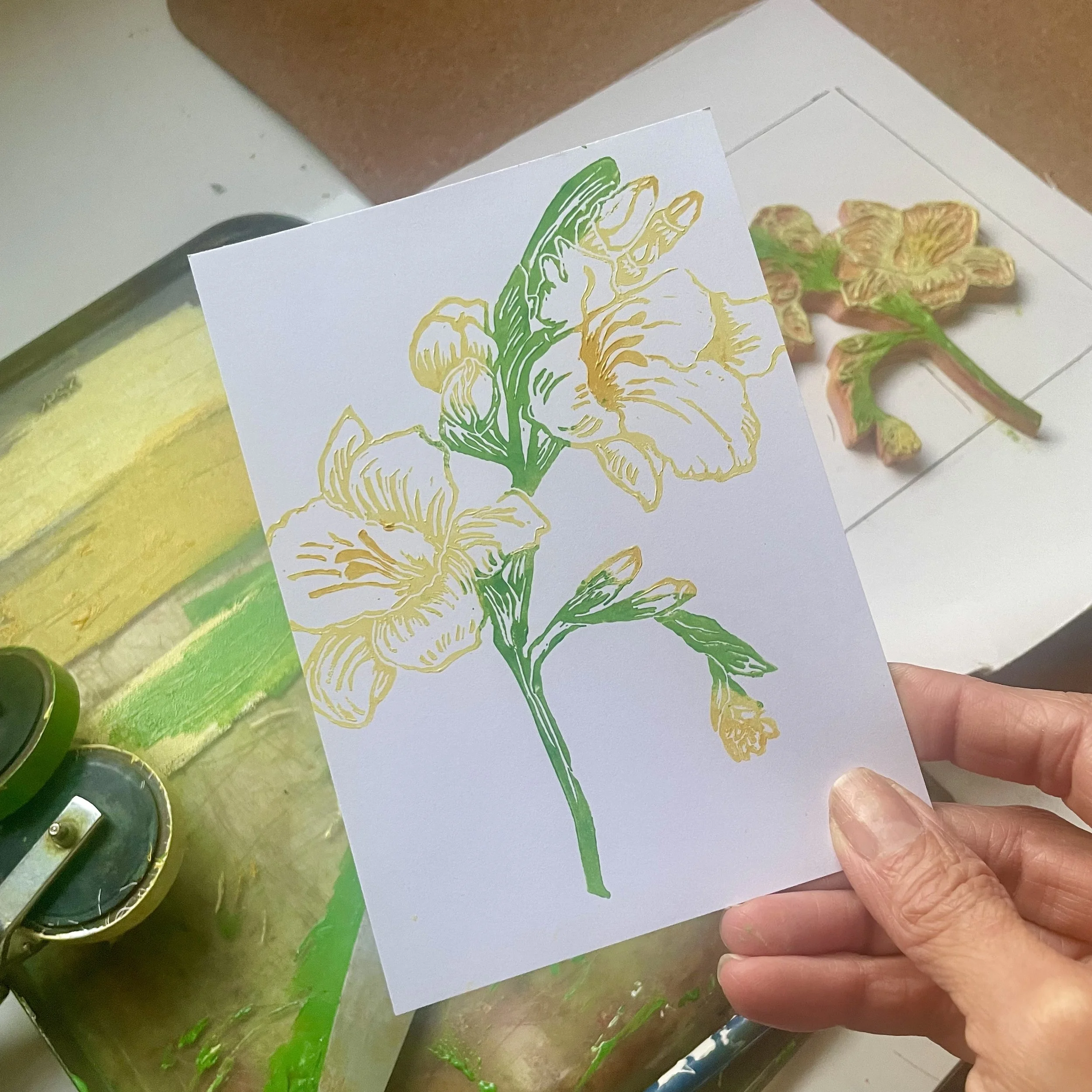 Hand holding a botanical print of yellow and green flower illustration, with a painted flower-shaped object in the background.