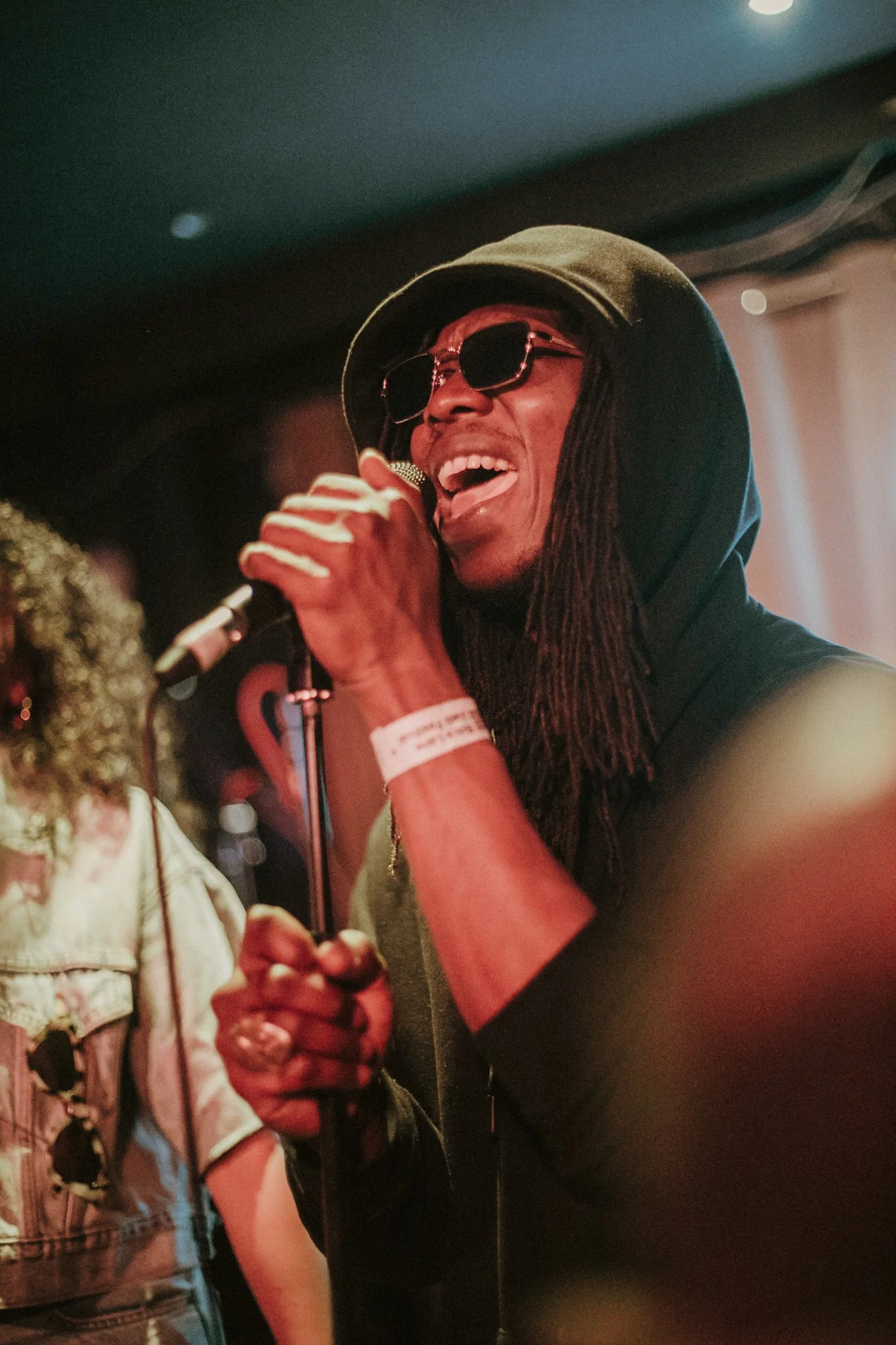 Person singing into a microphone, wearing sunglasses and a hoodie, in a dimly lit setting.