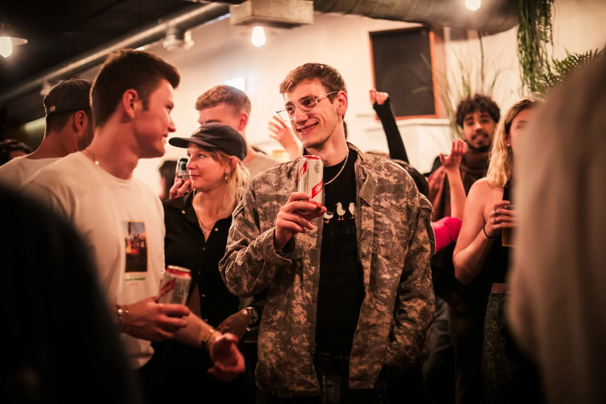 People socializing at a party, holding canned drinks, in a lively indoor setting with dim lighting and decorative plants.