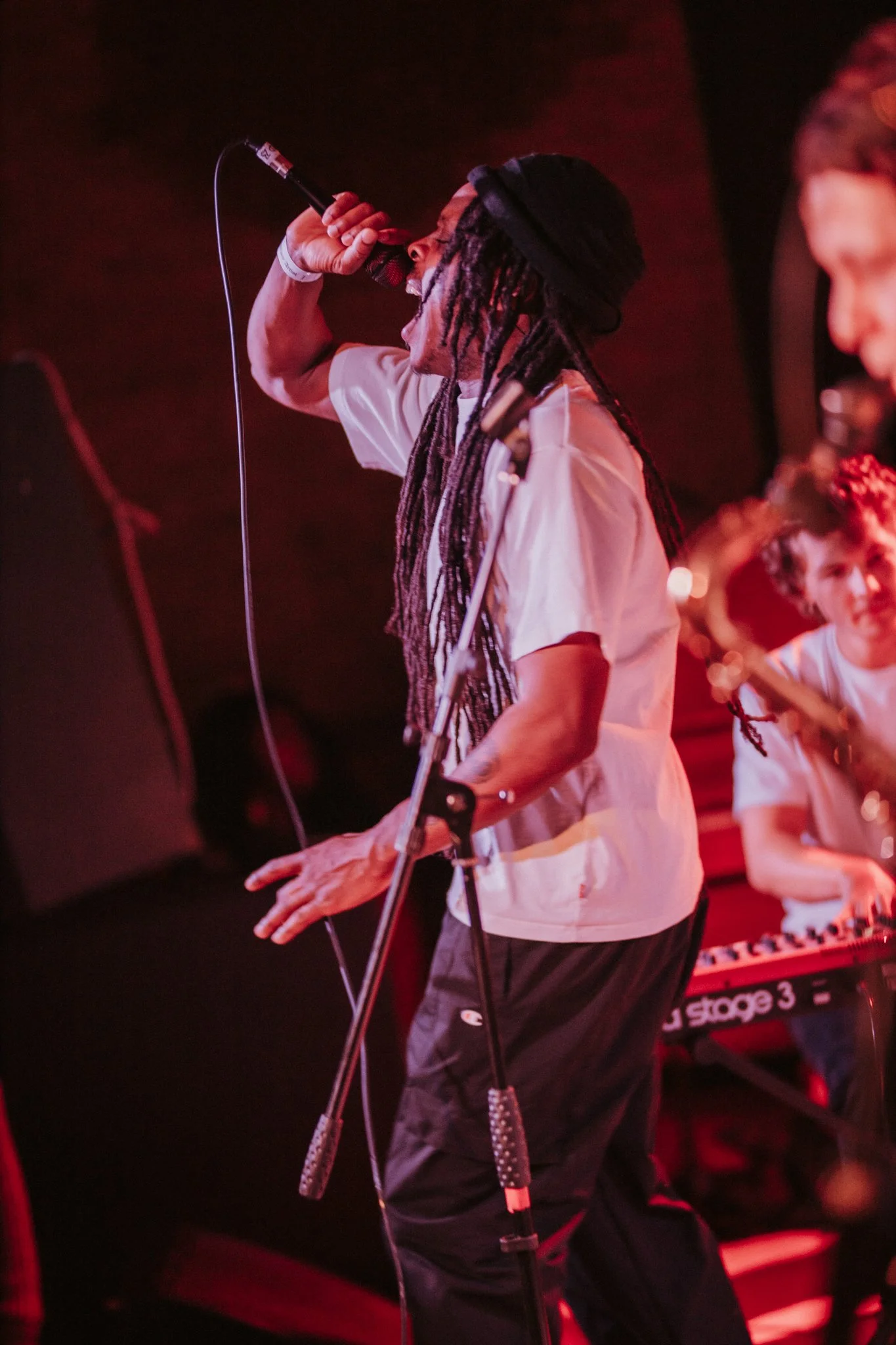 Musician singing passionately into a microphone on stage, with another musician playing keyboard in the background.