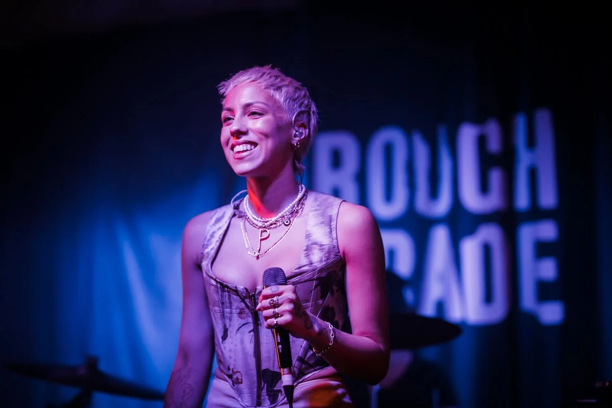 Person holding a microphone performing on stage with 'Rough Trade' sign in the background.