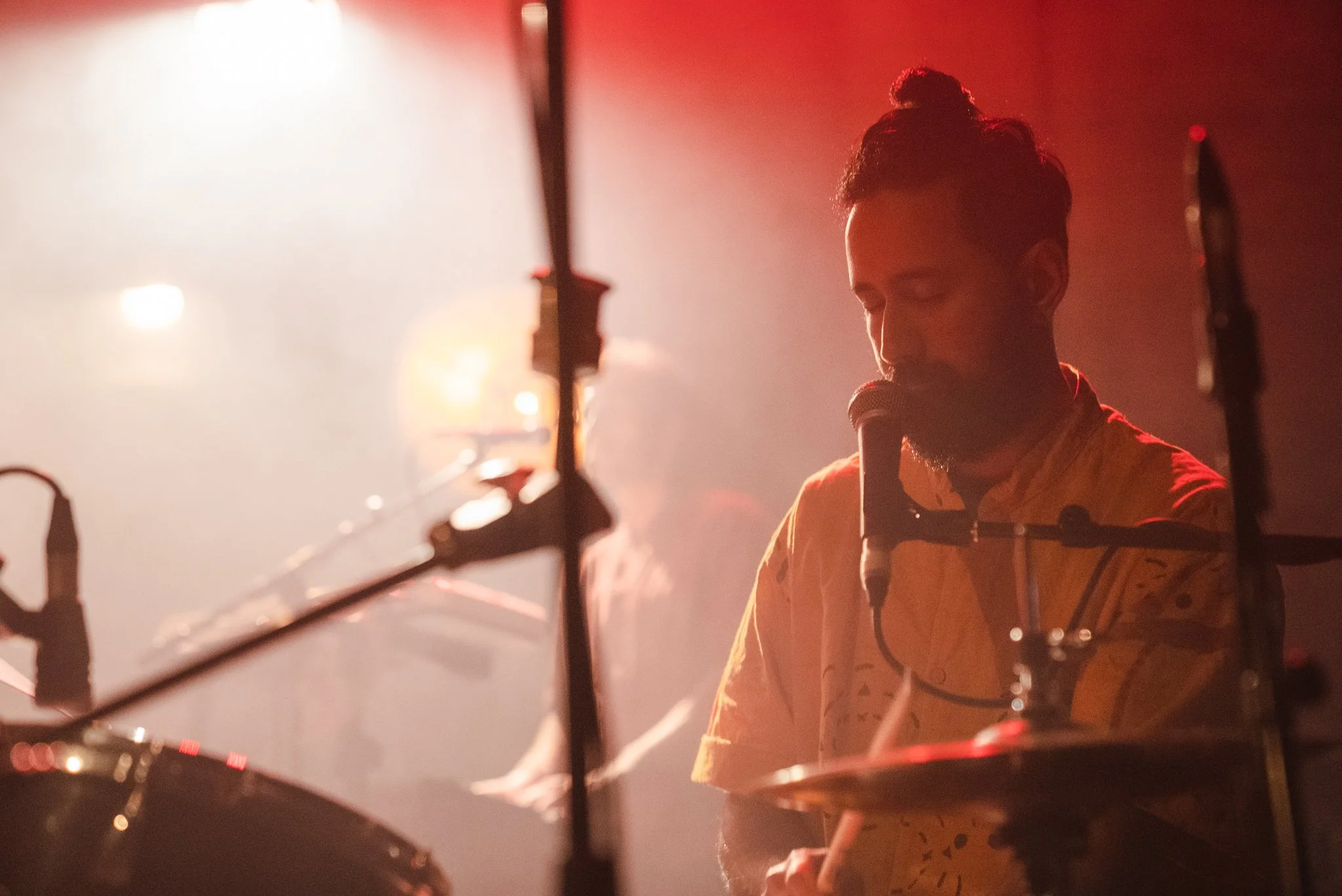 Musician playing drums and singing into a microphone on a dimly lit stage, with a red light backdrop.