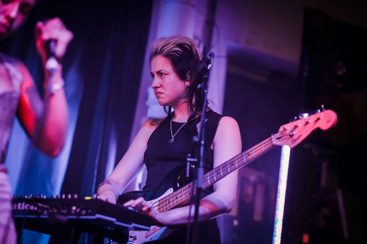 Musician playing bass guitar on stage with focused expression, wearing a sleeveless top, with neon lighting effects.