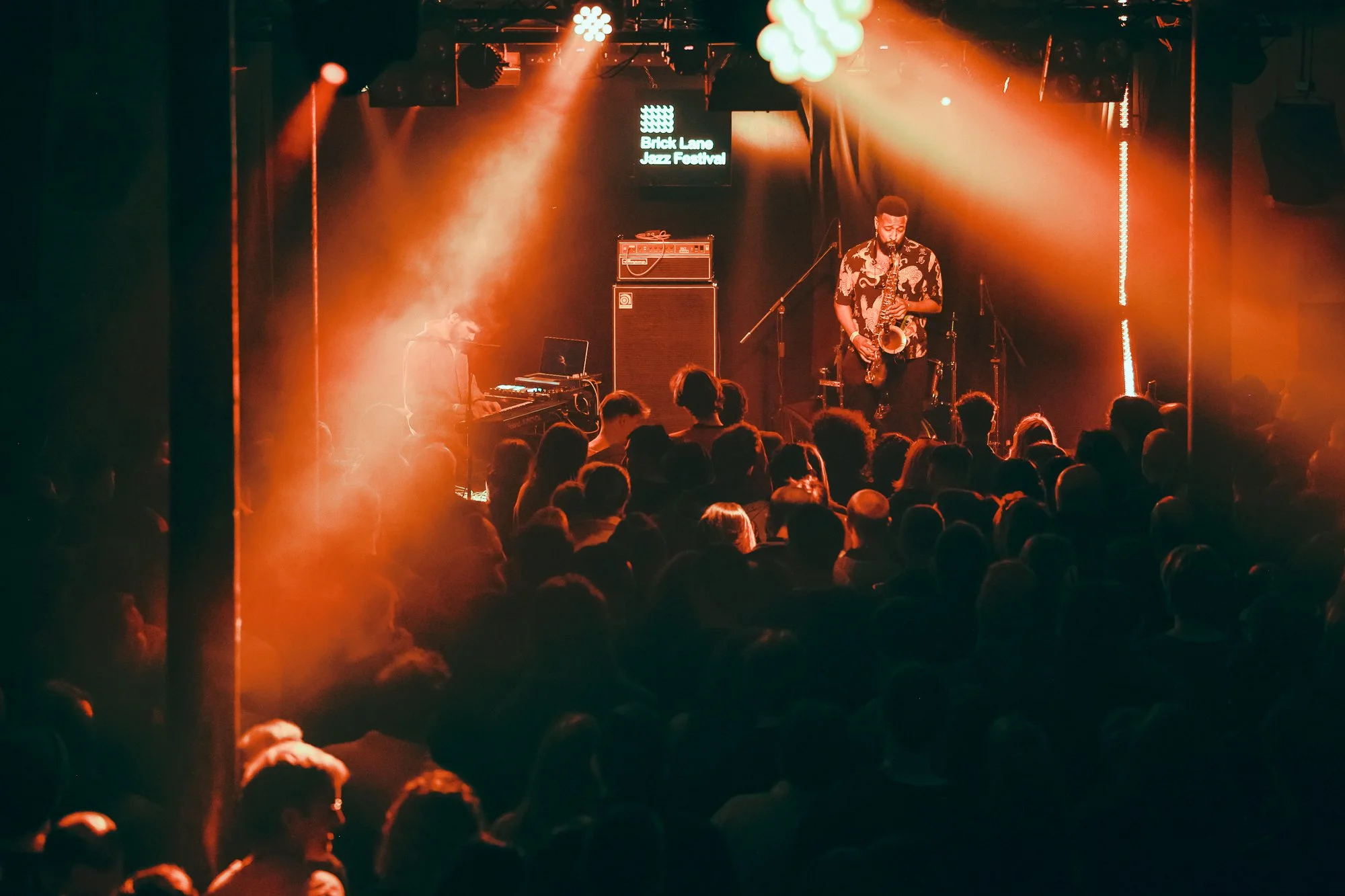 Musicians performing at Brick Lane Jazz Festival with audience in foreground and red lighting.