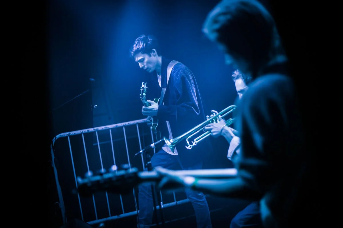 Live band performing on stage with blue lighting, featuring a guitarist and a trumpet player.