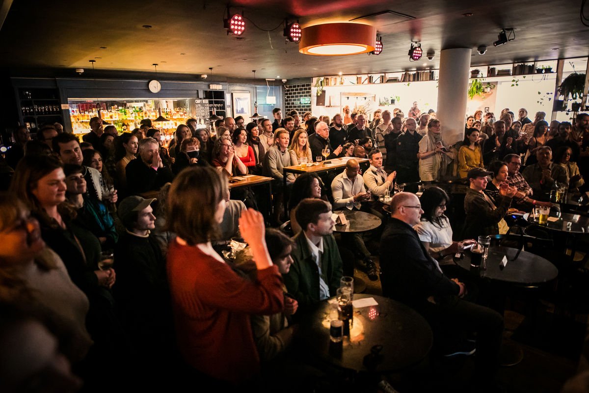 Crowded indoor event at a bar with audience seated and standing, people holding drinks, attentive to a presentation or performance.