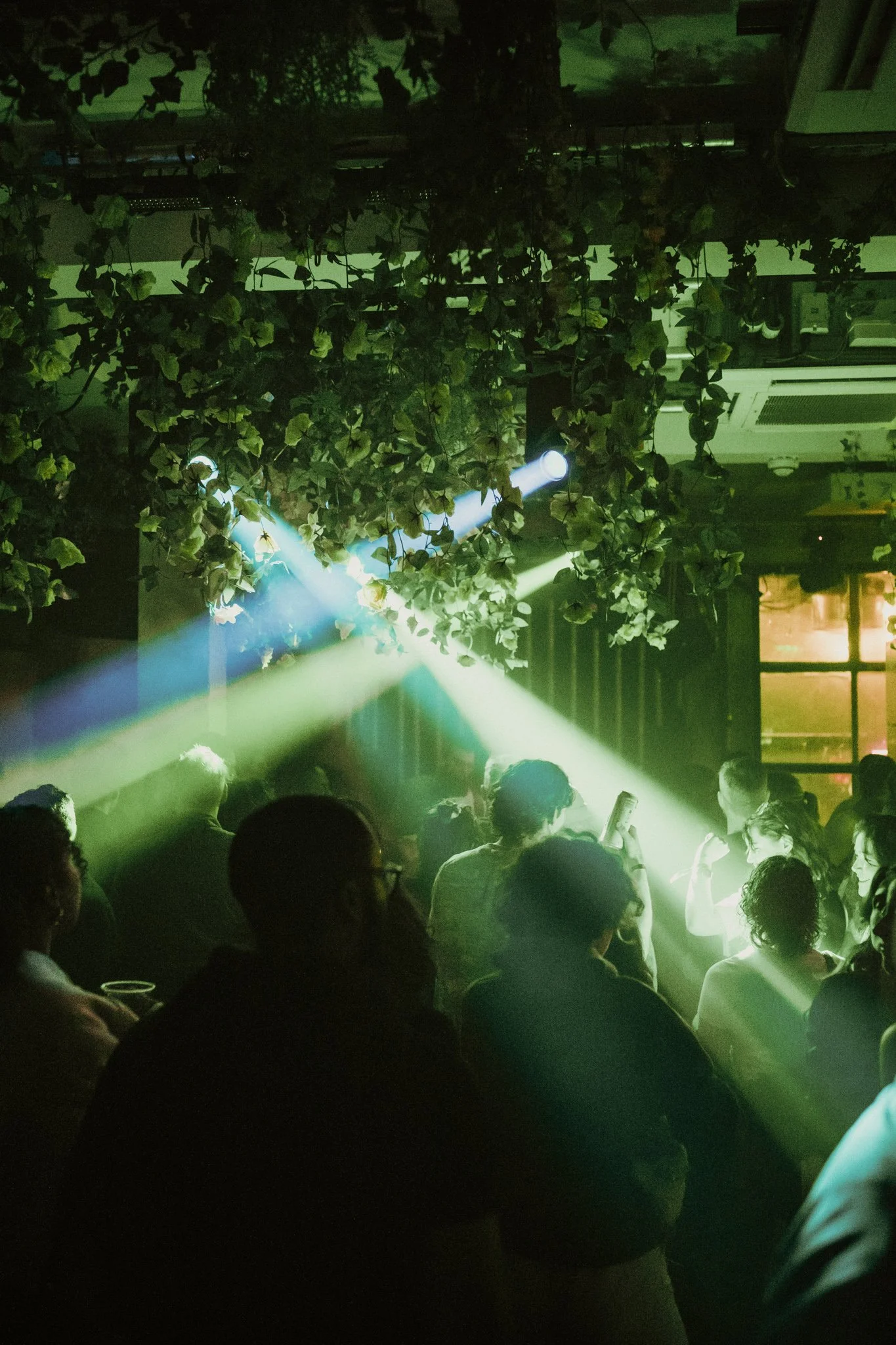 Nightclub scene with green lighting, plants hanging from the ceiling, and a crowd of people dancing.