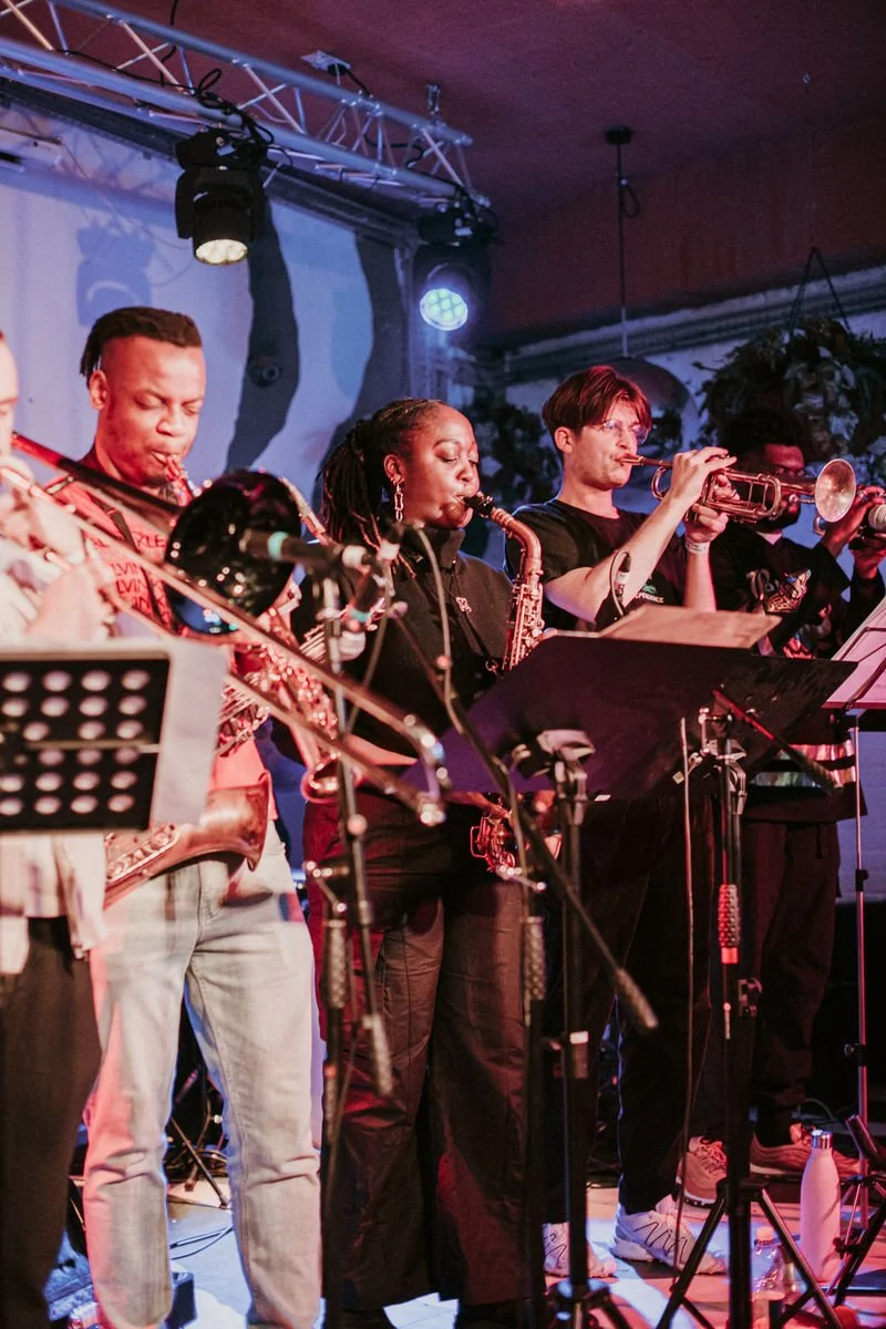 A group of musicians performing on stage with brass instruments, including saxophones and trumpets, amid a colorful lighting setup.