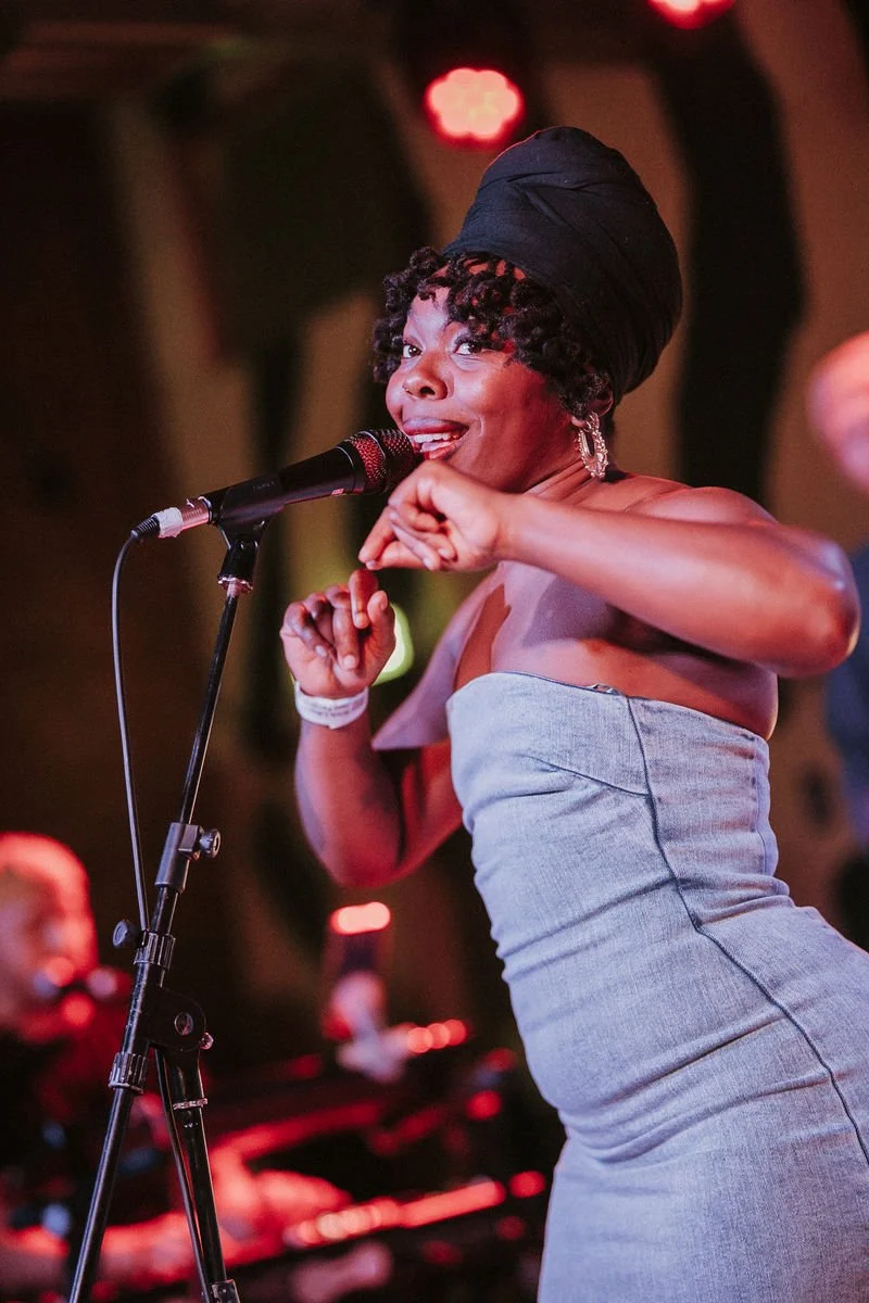 Singer performing on stage with microphone, wearing a strapless dress and headwrap.