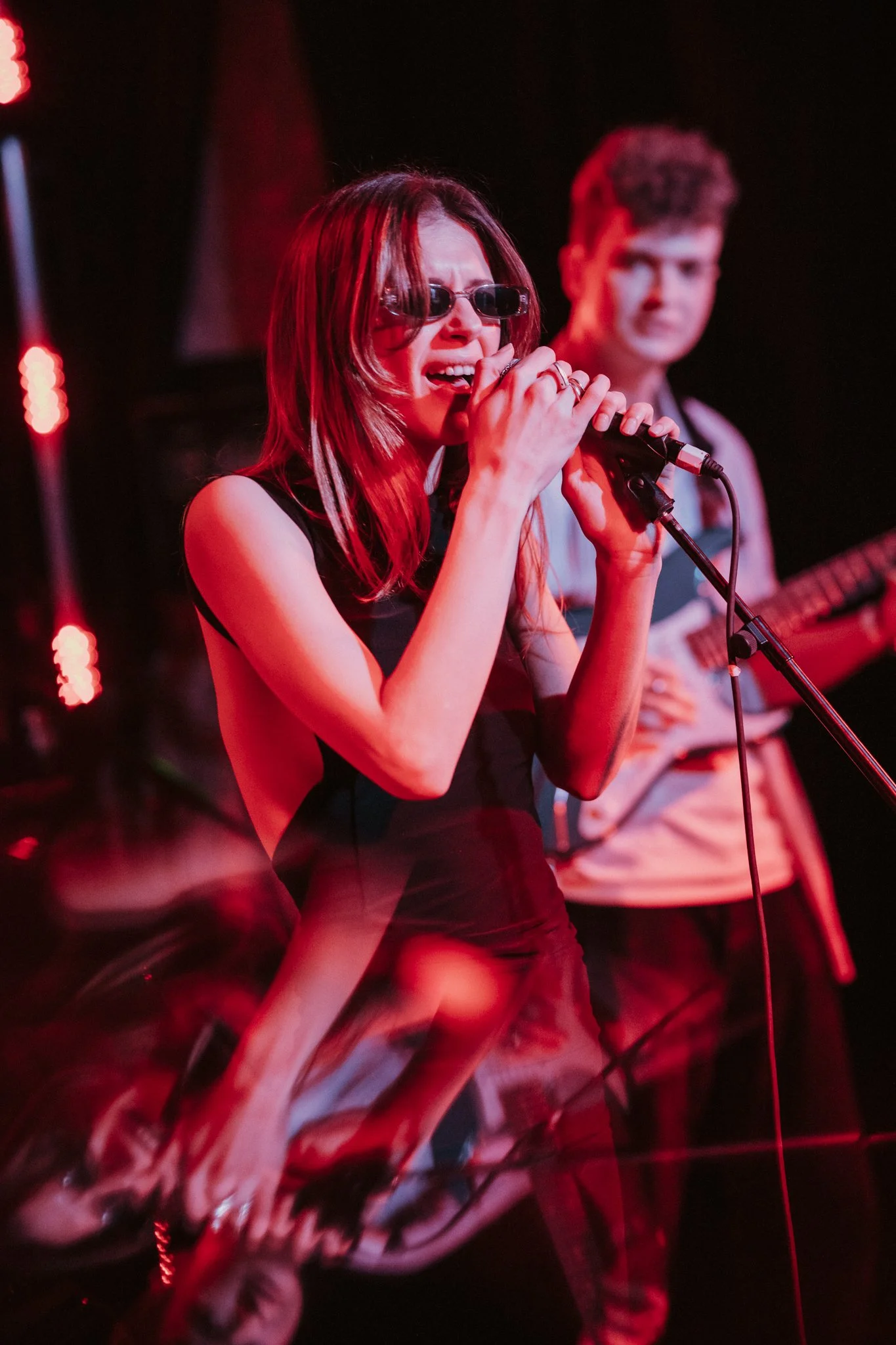 Singer on stage with microphone and guitarist in background, red lighting.