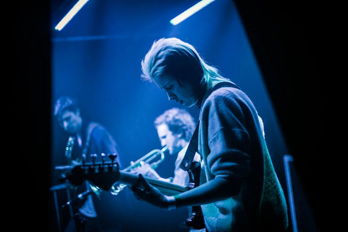 Musicians performing on stage with blue lighting, featuring a guitarist in the foreground and a trumpet player in the background.