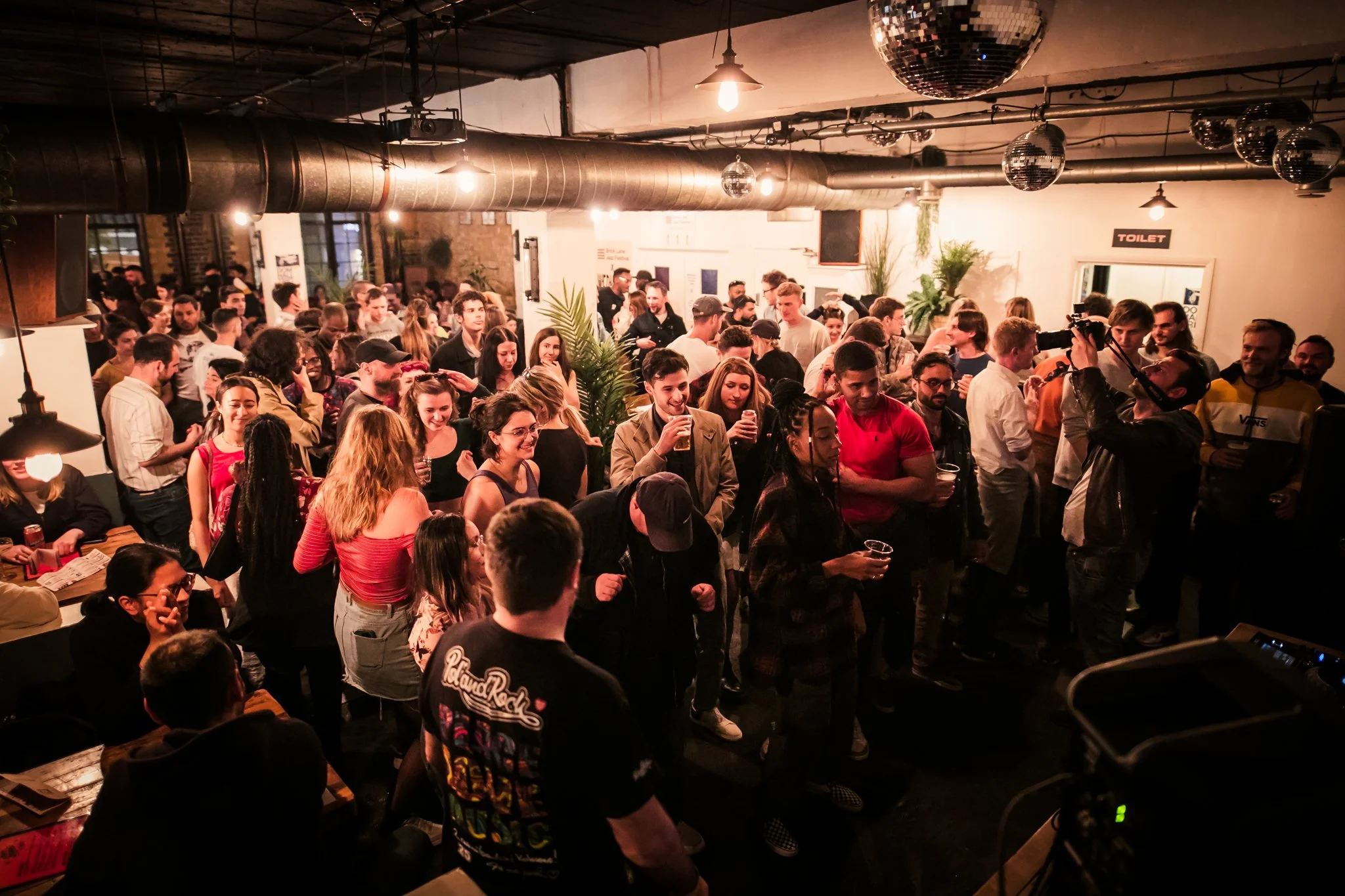 Crowded party scene in a dimly lit venue with people socializing and dancing. Ceiling features exposed pipes and disco balls.