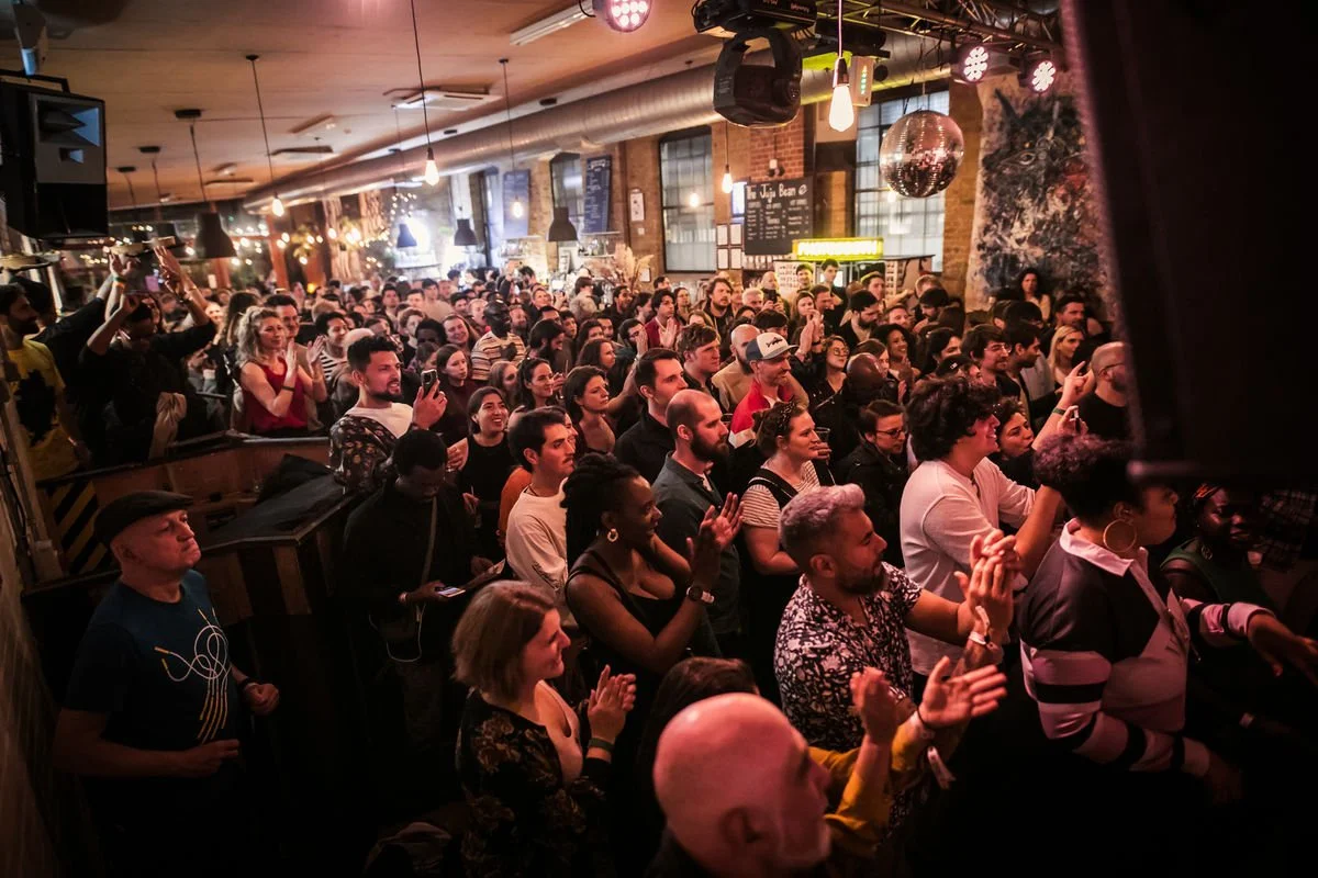 Large crowd in a lively indoor venue, people clapping and smiling, dim lighting with hanging lights and a disco ball.