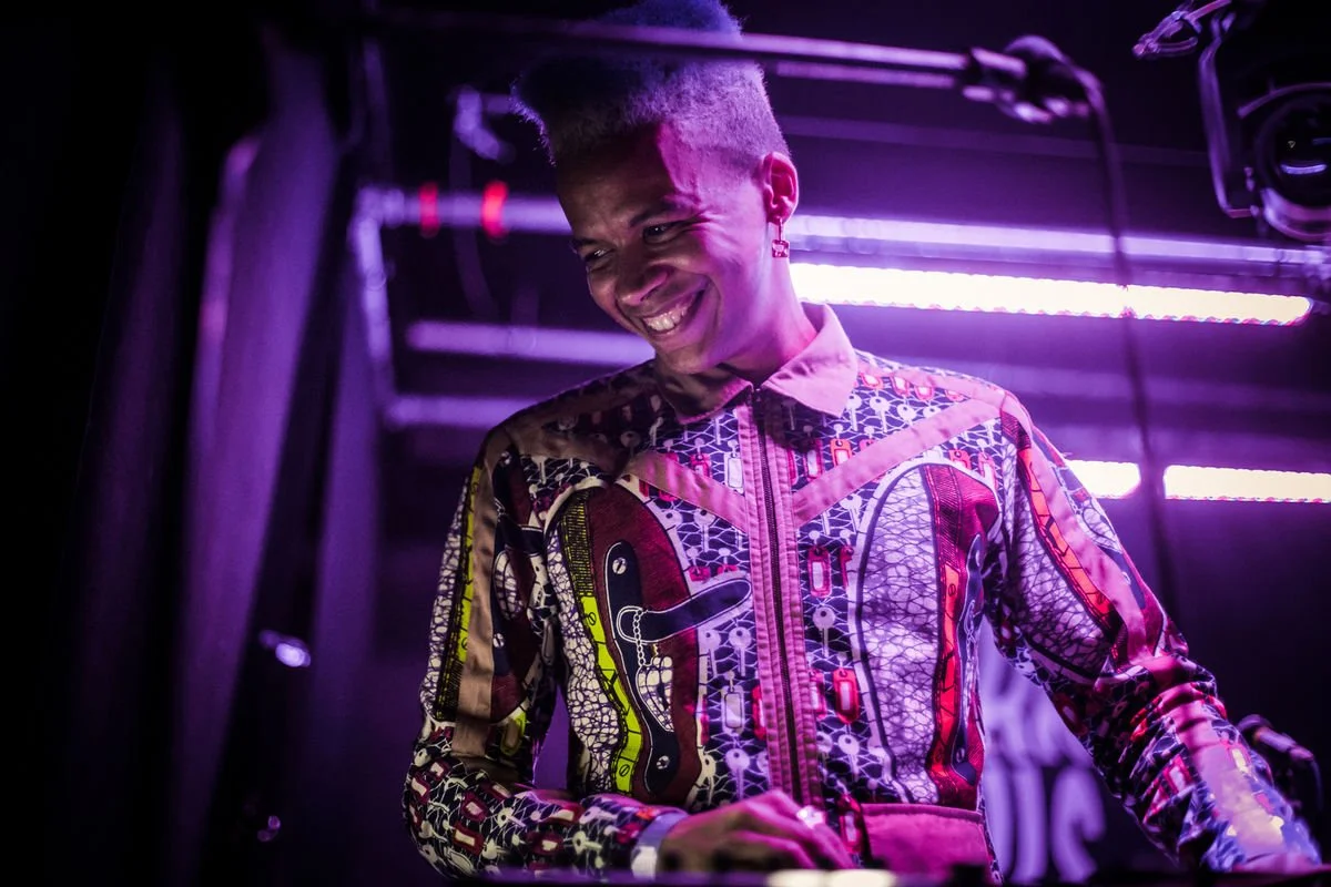 Person in a colorful, patterned outfit smiling on stage with purple lighting.
