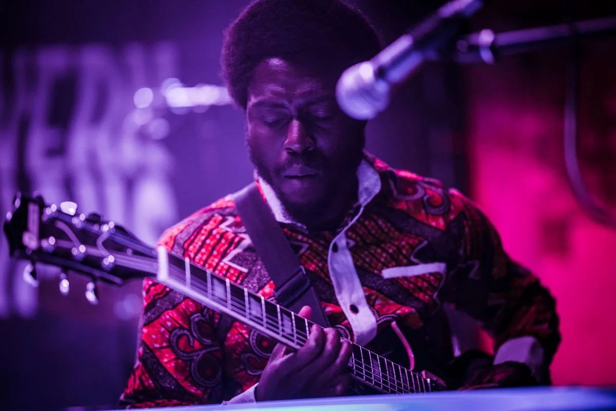 Musician playing guitar on stage with microphone, wearing colorful patterned shirt, purple lighting.