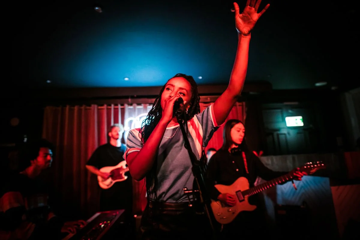 Band performing on stage with vocalist, guitarists, and keyboardist under red and blue lights.