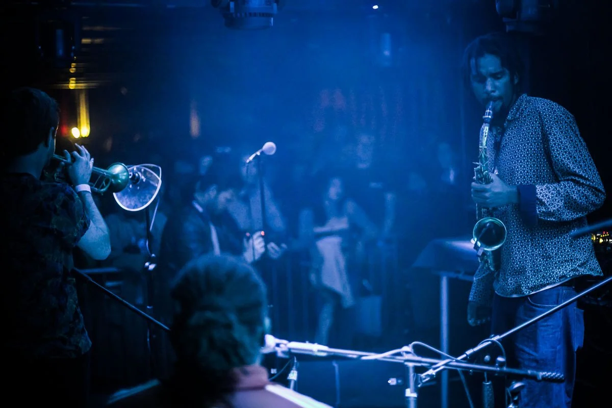 Musicians playing trumpet and saxophone in a dimly lit venue with an audience in the background.