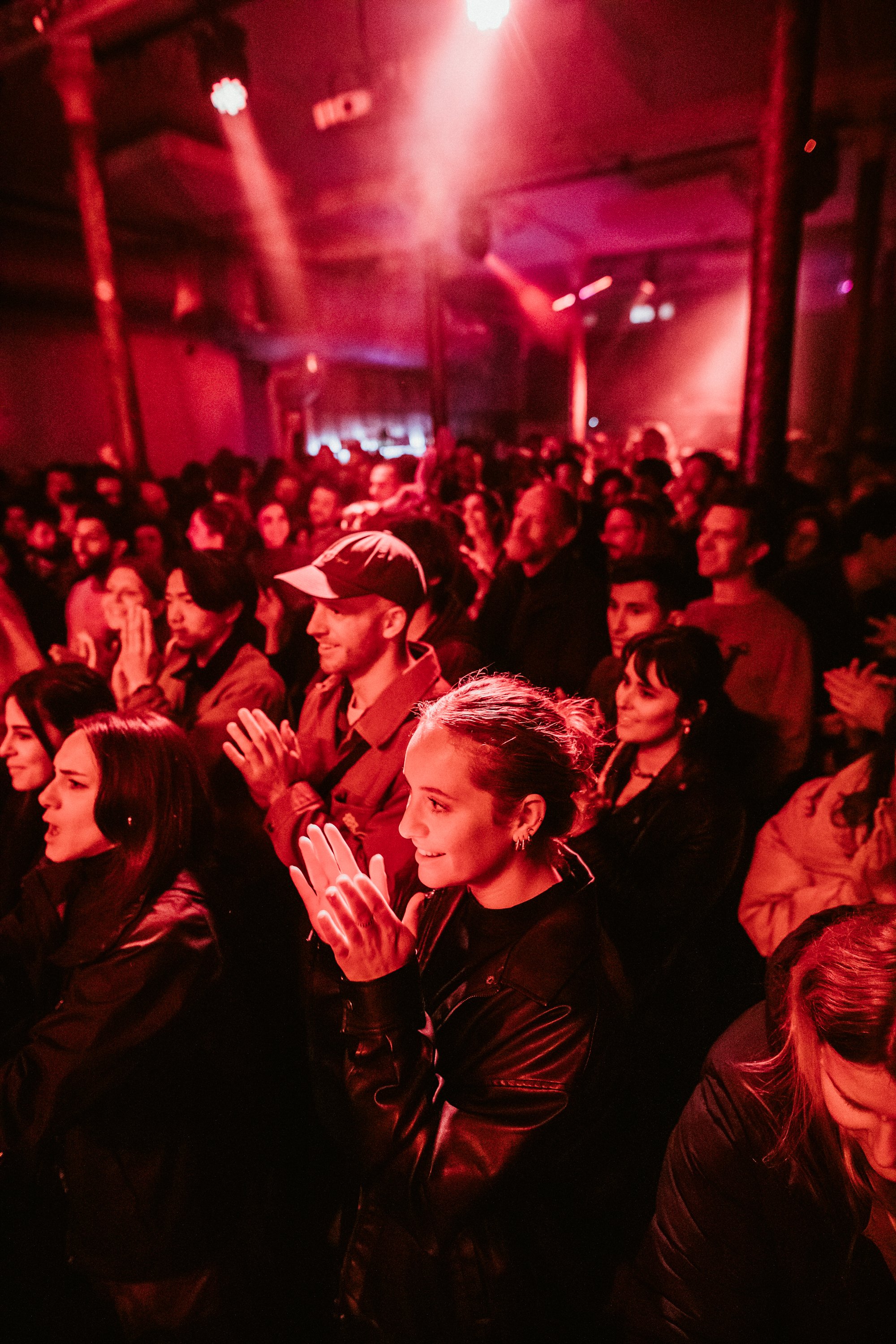 Crowd at a concert under red lighting, people clapping and enjoying the performance.