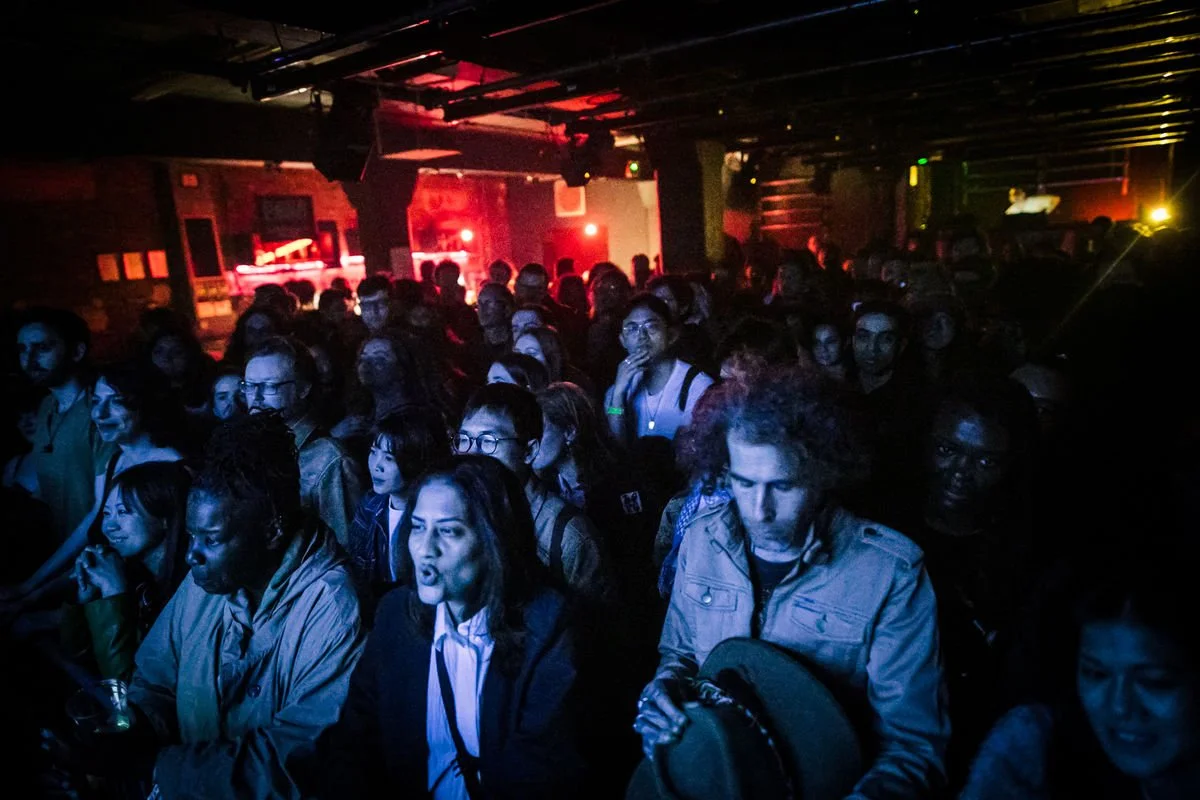 Crowd enjoying a live music concert in a dimly lit indoor venue with red and blue lighting.