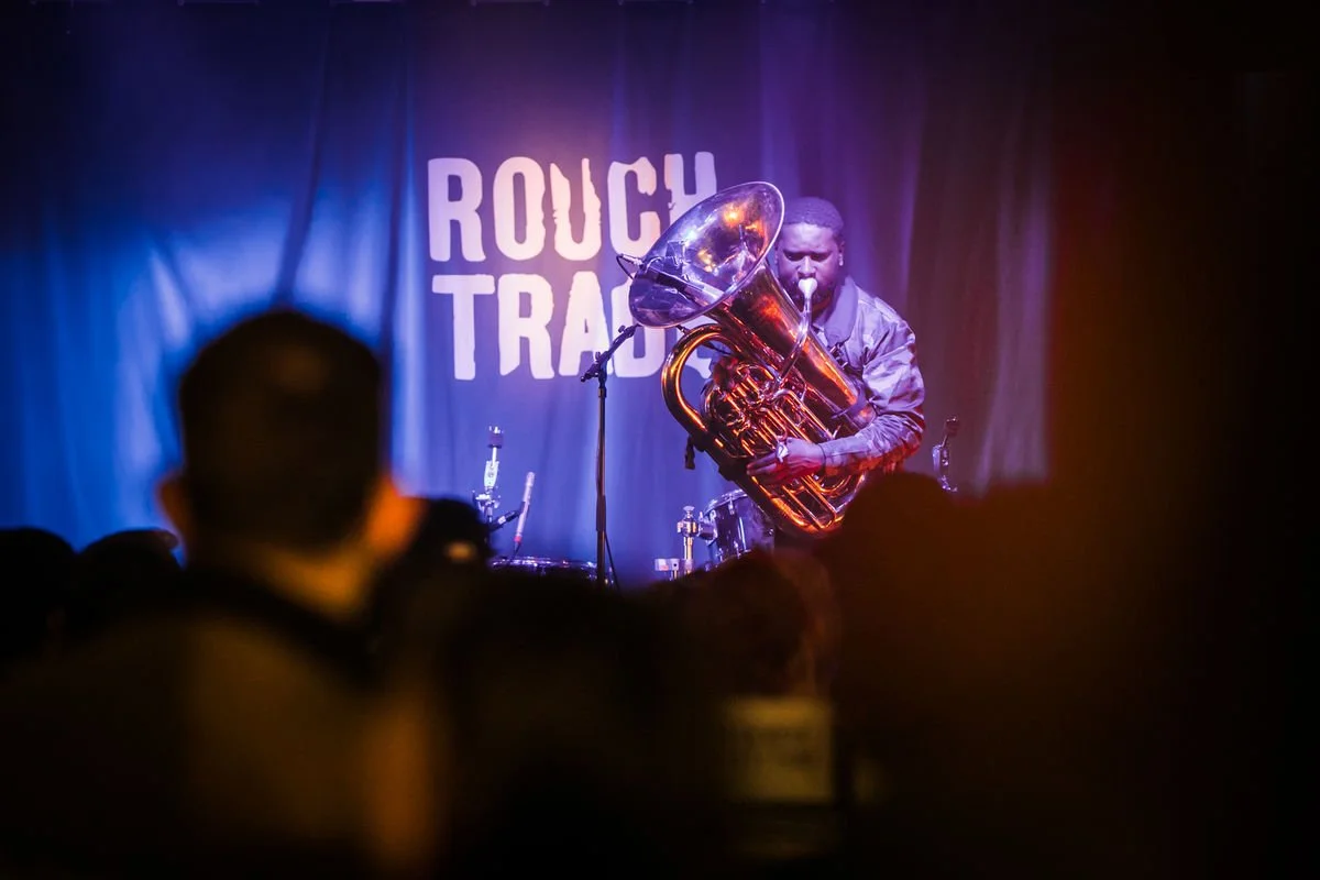 Musician playing a tuba on stage at Rough Trade event with audience in foreground.