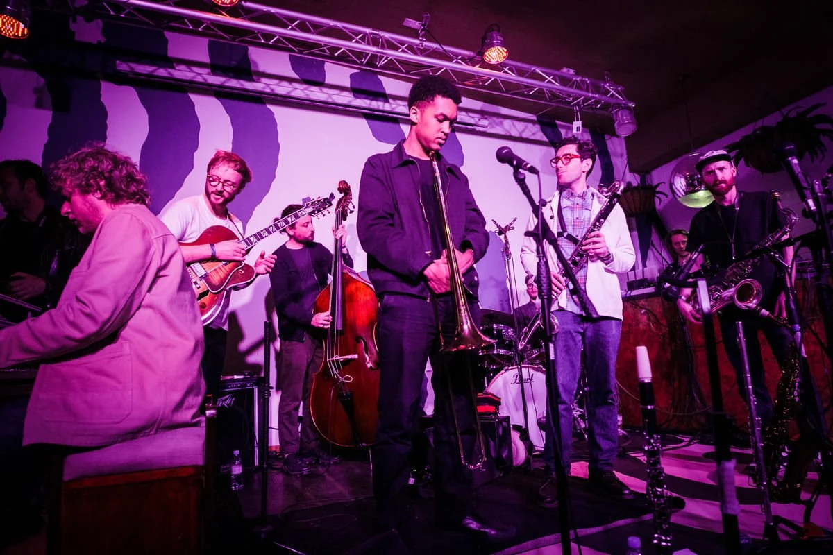 Live jazz band performing, featuring musicians with a trombone, electric guitar, double bass, clarinet, and saxophones on a dimly lit stage.
