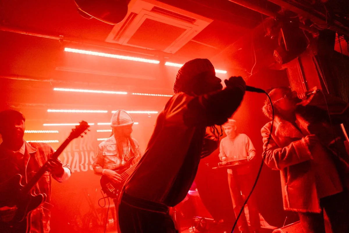 Musicians performing on stage with red lighting, featuring guitarists, vocalists, and a DJ in a club setting.